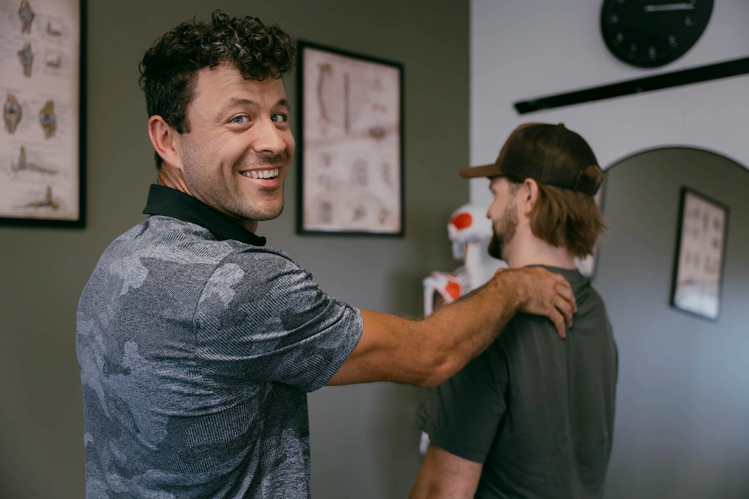 A man with curly hair smiling and putting his arm around another person in a room with medical posters and a skull model.