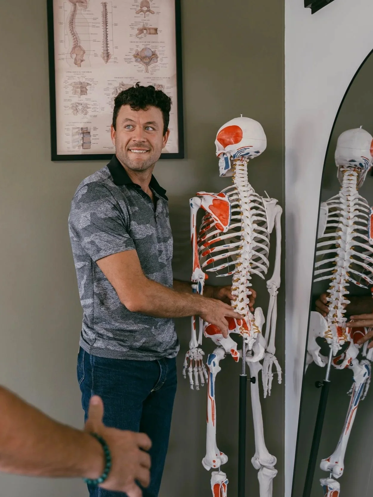 A man standing next to a skeleton model with muscles painted on it, in front of a mirror. There is a medical diagram on the wall behind him.