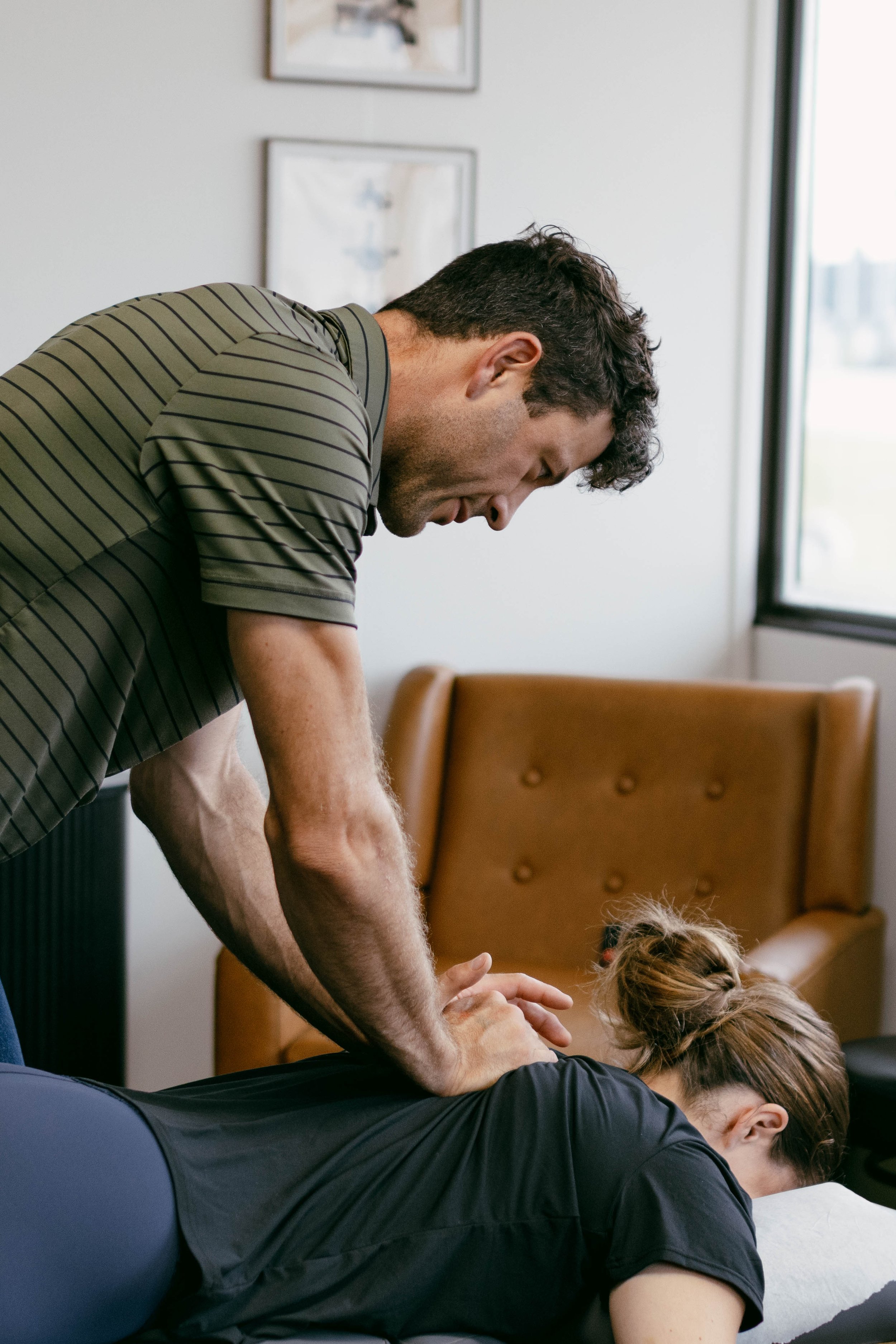 A man giving a massage to a woman lying face down on a massage table in a room with framed pictures on the wall and a window letting in natural light.