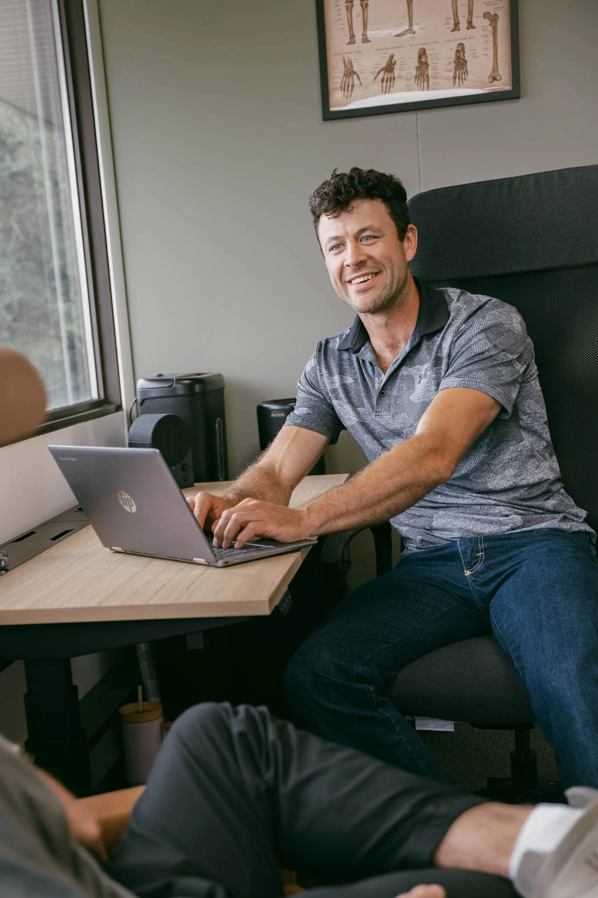 Man sitting at a desk using a laptop computer in an office, with medical posters of bones on the wall behind him.