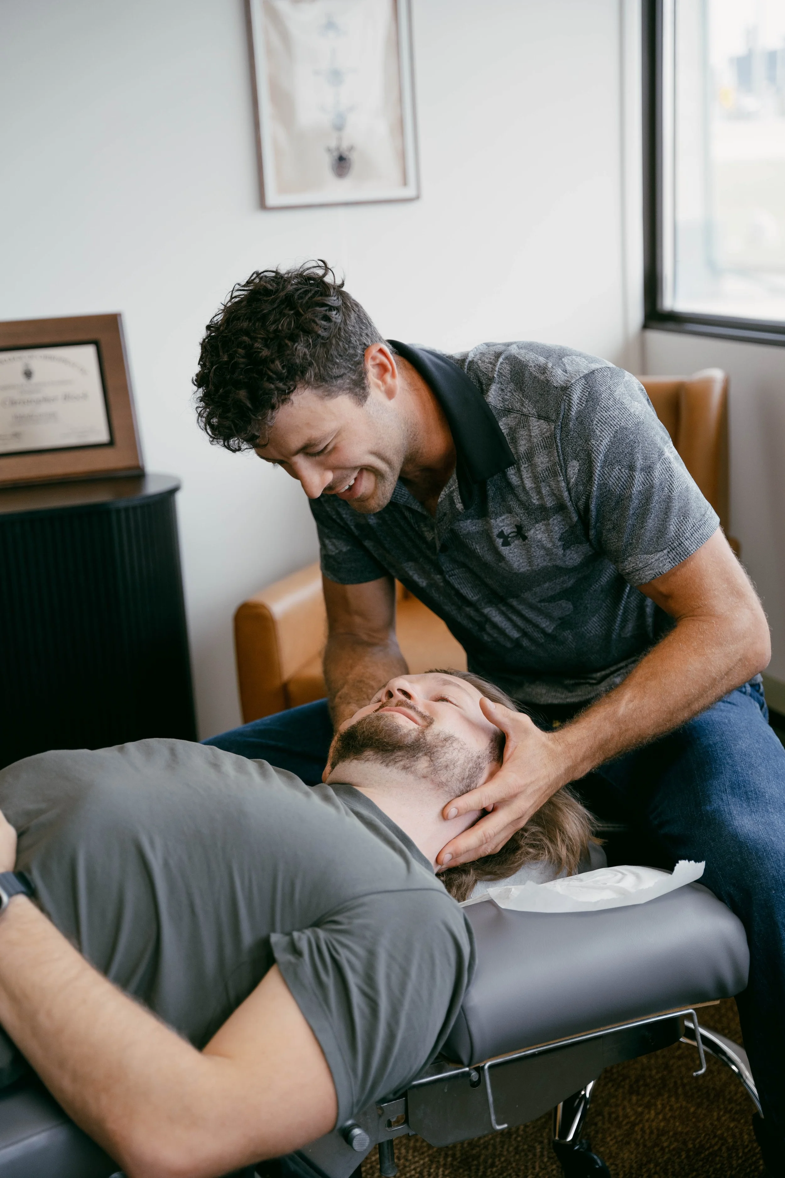 A man with long hair is lying on a medical examination table with his eyes closed, while another man is performing manual CPR or rescue breathing, holding his head and neck and looking down at him in a well-lit room with a window and medical certificates or diplomas on the wall.