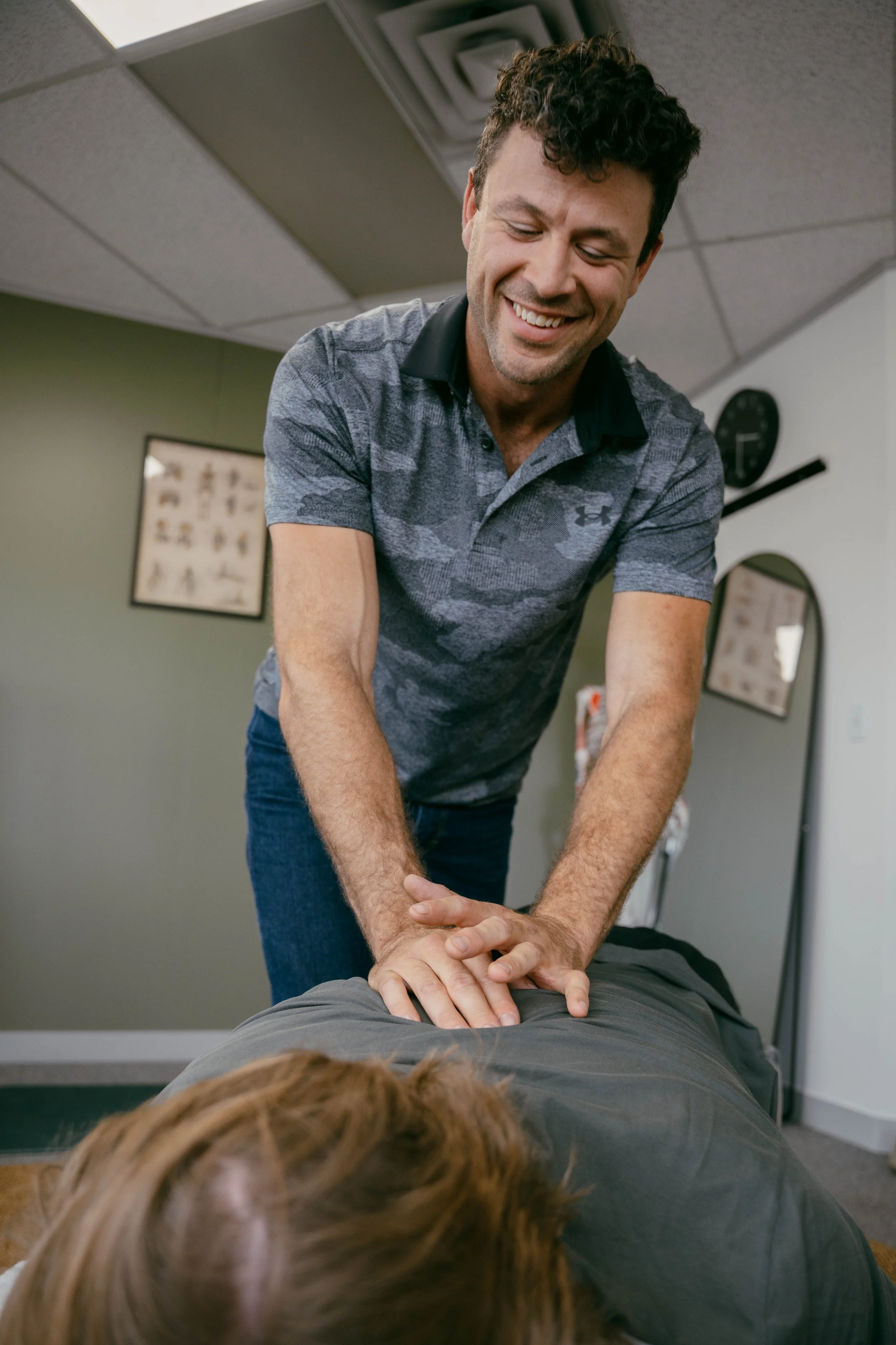 A chiropractor or physical therapist performing an adjustment or massage on a person lying face down on a massage table, with the therapist smiling and the patient holding onto the table.