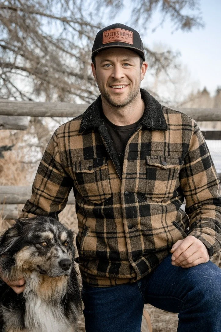 A smiling man in a plaid jacket and baseball cap kneeling next to an Australian Shepherd dog outdoors with a wooden fence and trees in the background.