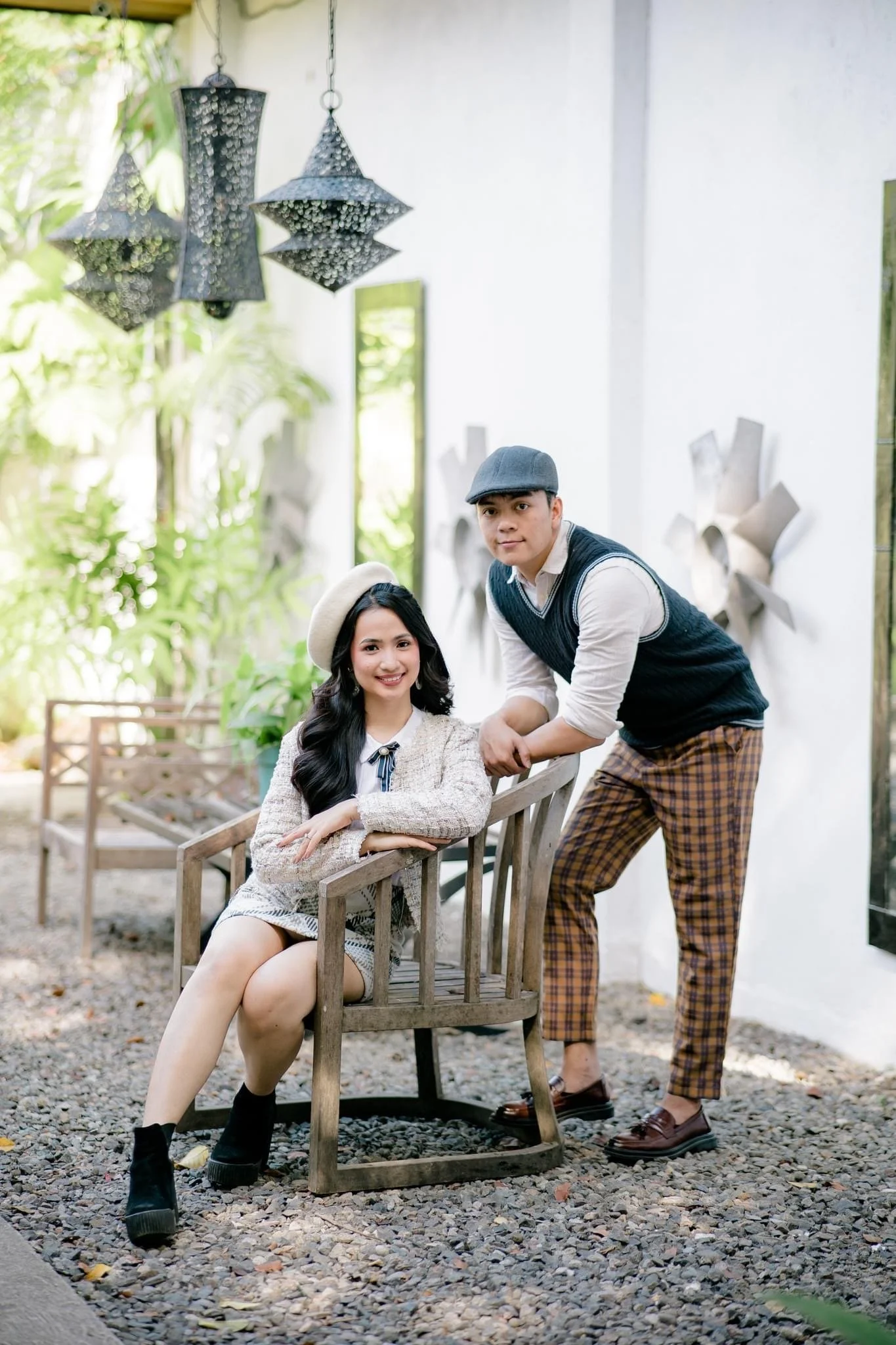 A young woman with long black hair, wearing a beige hat, patterned dress, and black boots, is sitting on a wooden bench. A young man with short hair, wearing a gray hat, white shirt with a blue vest, and plaid pants, is standing beside her, holding her hand. They are outdoors, surrounded by greenery and modern wall art, with hanging decorative lamps above them.