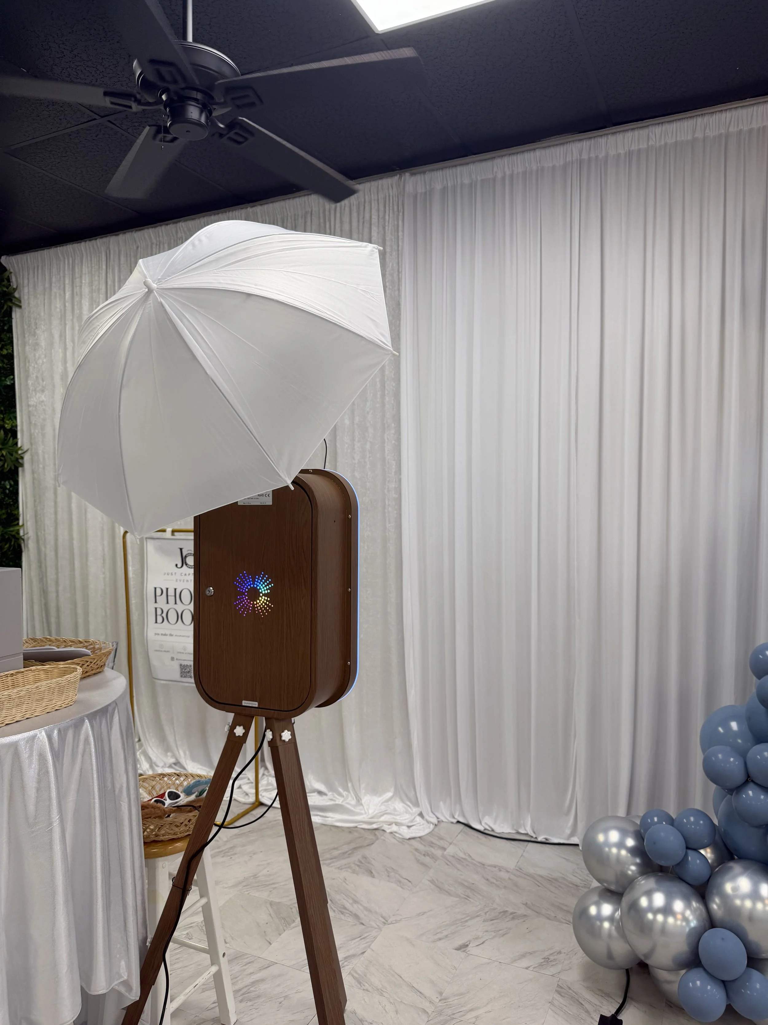 Indoor photo of photo booth setup with a white umbrella, a wooden photo booth frame, and a backdrop with white curtains. Decor includes a cluster of blue and silver balloons on the right side.