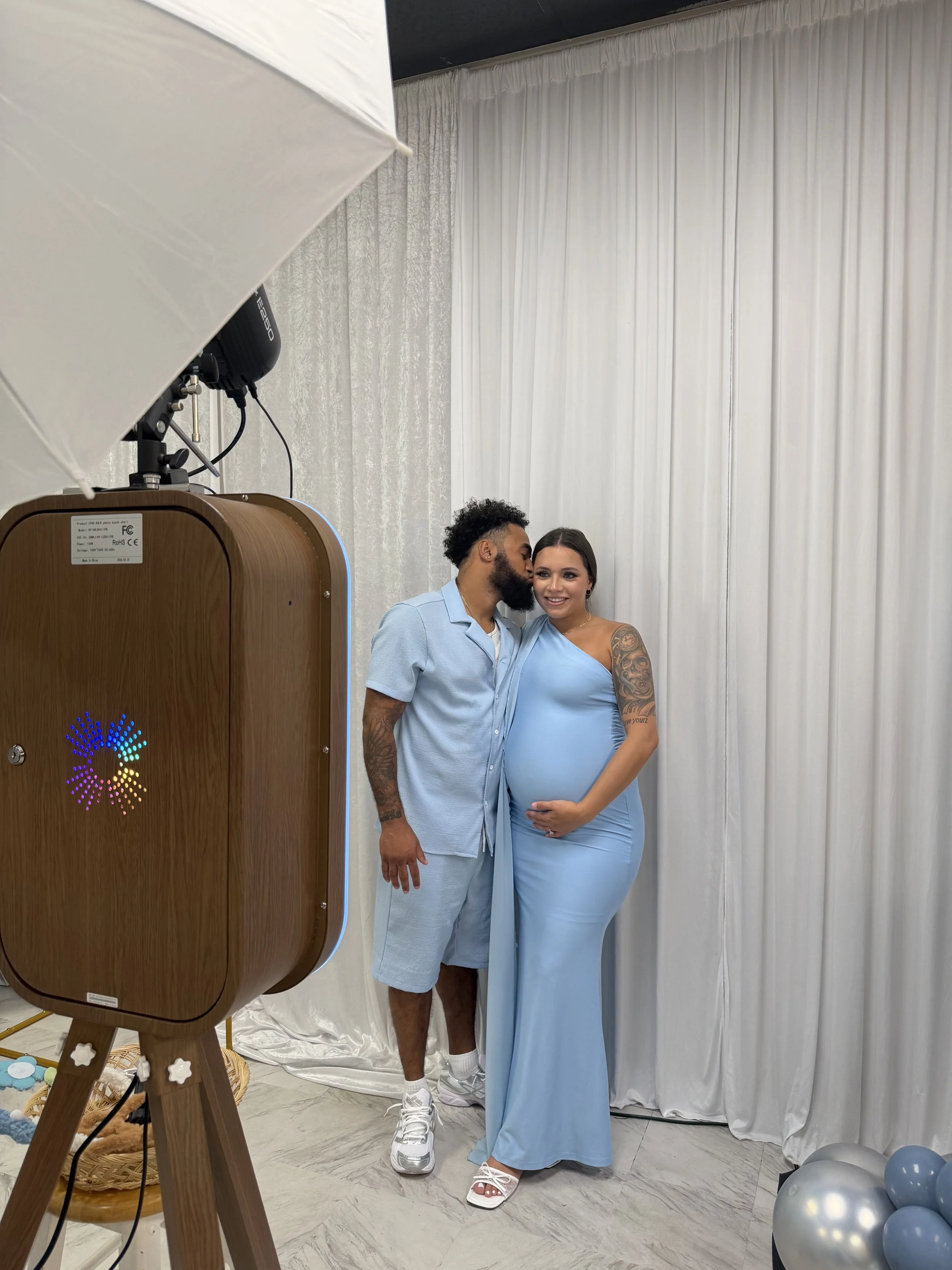 A pregnant woman with long dark hair wearing a light blue dress and white sandals standing next to a man with a beard and curly hair wearing a light blue shirt and shorts, with balloons on the floor nearby, in front of a white curtain backdrop at a celebration.