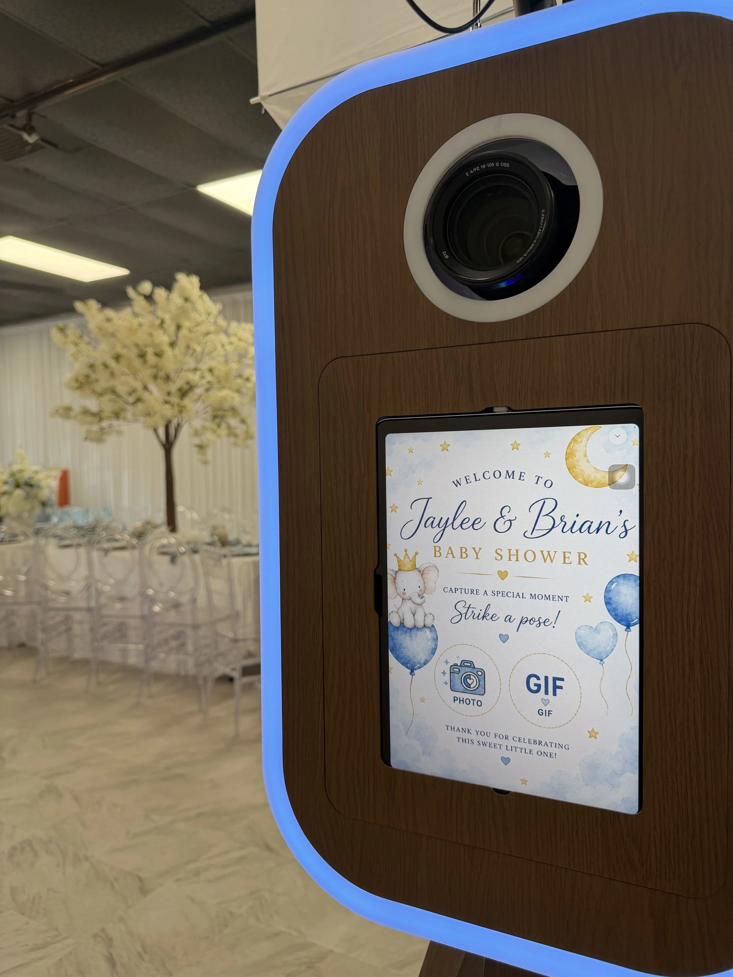 Photo of a digital screen on a wooden stand Photo Booth at a baby shower, with a welcome message and options to share photos or GIFs. The background shows tables set for a celebration with chairs and floral arrangements.