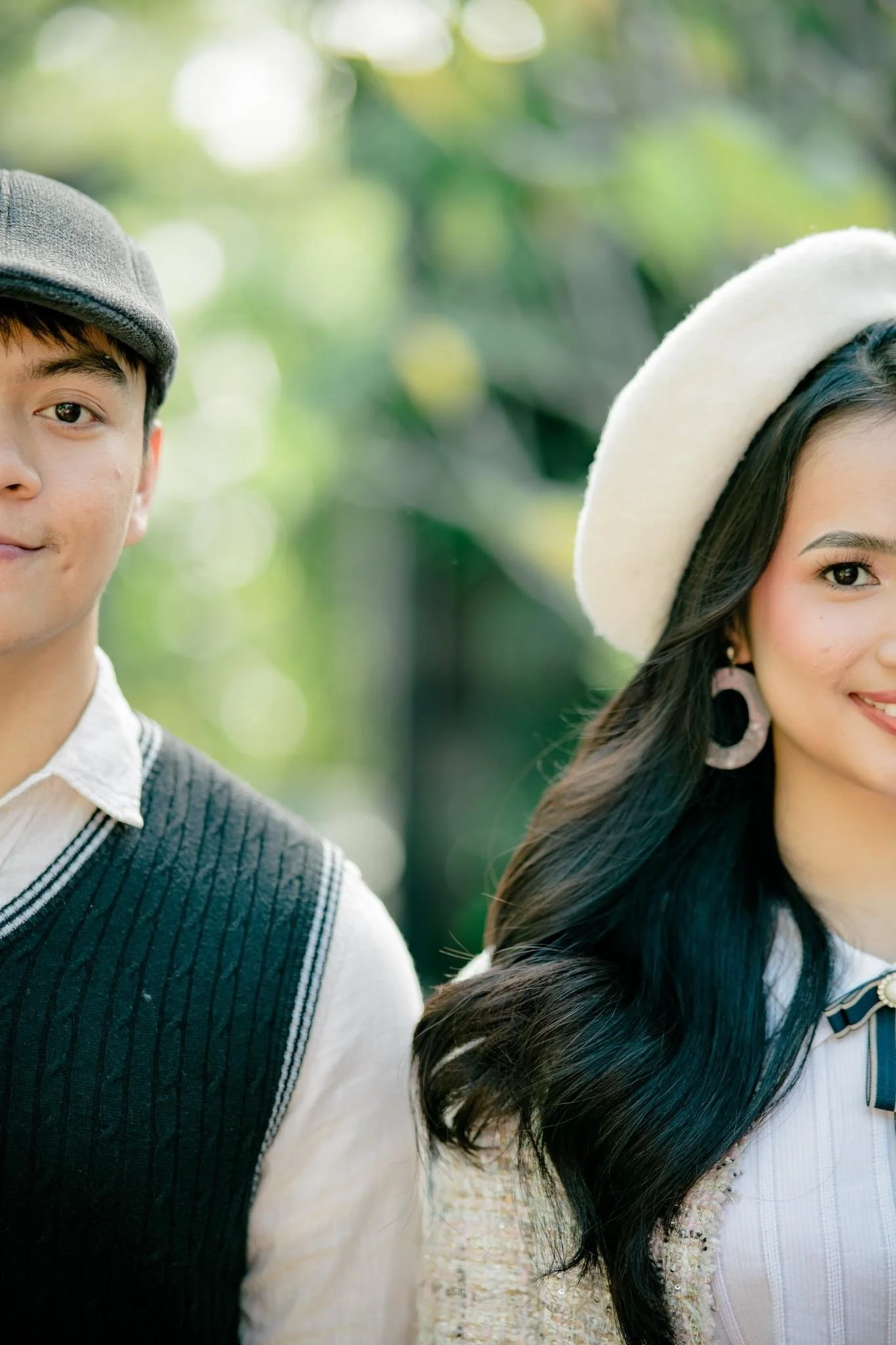 Close-up of a man on the left and a woman on the right in an outdoor setting with greenery, wearing hats and smiling.