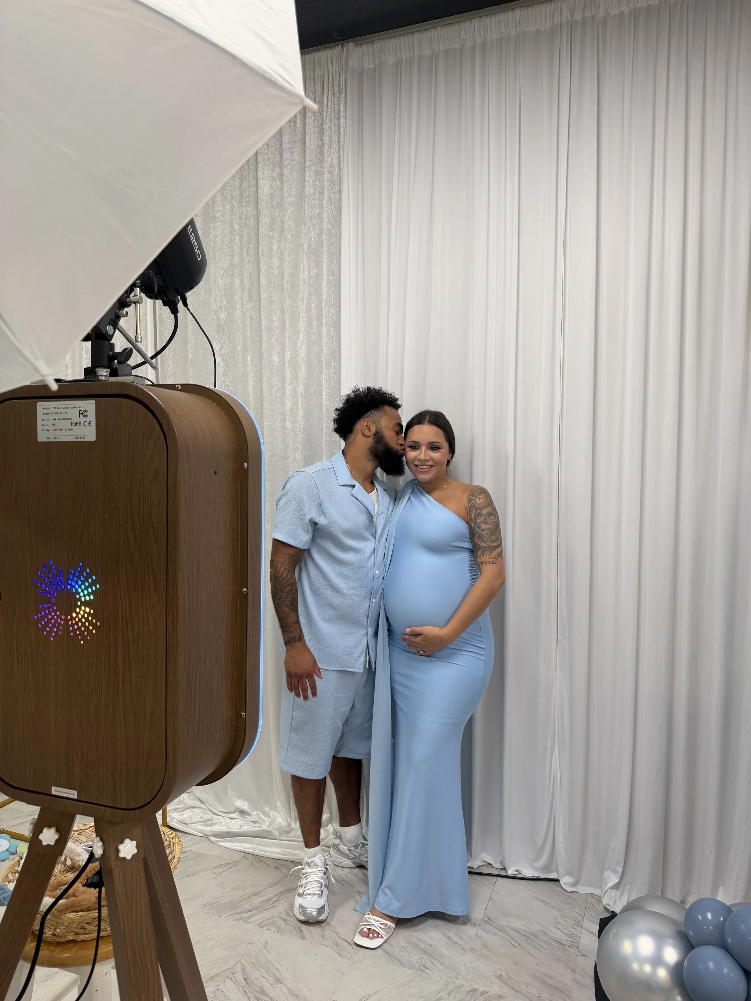 A couple takes a photo in a photo booth, the woman is pregnant and is wearing a light blue dress while the man is wearing a light blue shirt and shorts. There is a large white curtain backdrop and some balloons on the floor.
