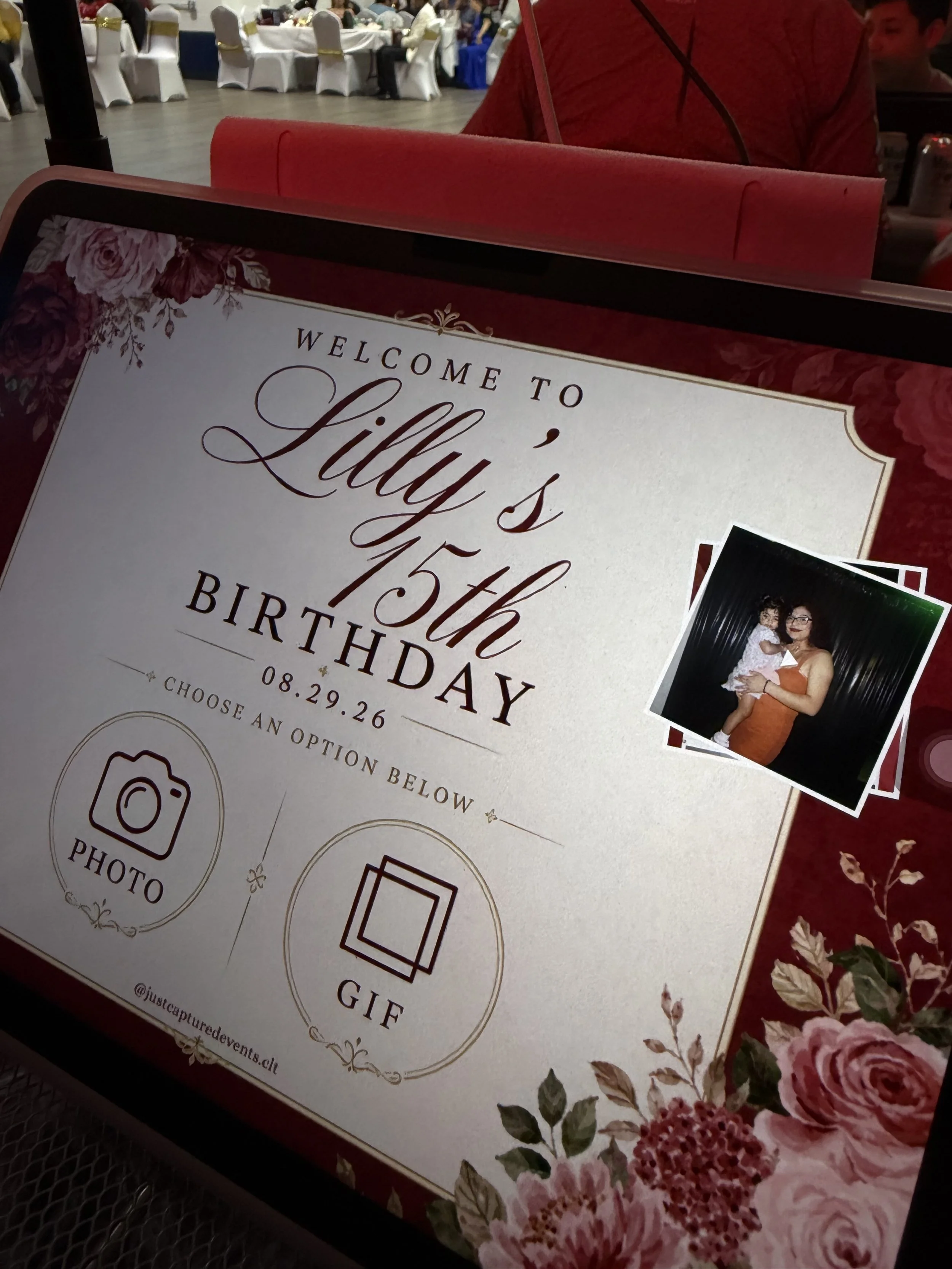 A sign with floral decorations welcomes guests to Lily's 15th birthday celebration at a banquet hall. The sign features options for taking photos, GIFs, and gifts. A woman holding a young girl is visible in a photograph attached to the sign. The background shows round tables with white chairs in the decorated venue.