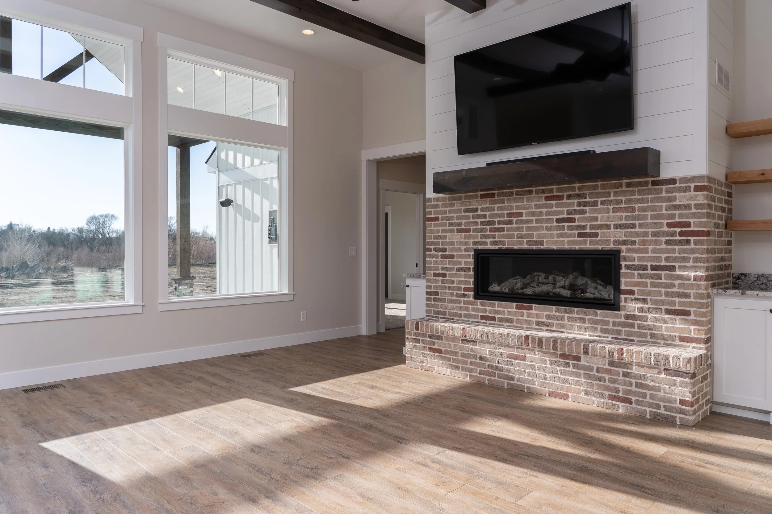 Living room with large windows, a brick fireplace and wall-mounted TV at JAB Construction’s Country Craftsman custom home in Shelley, Idaho.