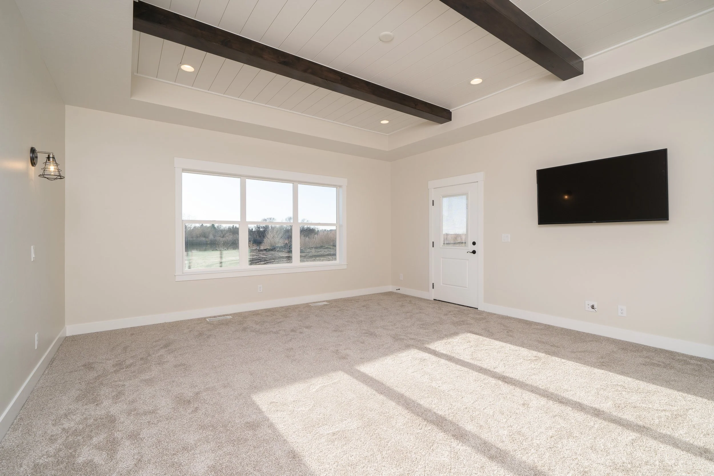 Carpeted living room with a large window, wall-mounted TV and ceiling beams at JAB Construction’s Country Craftsman custom home in Shelley, Idaho.