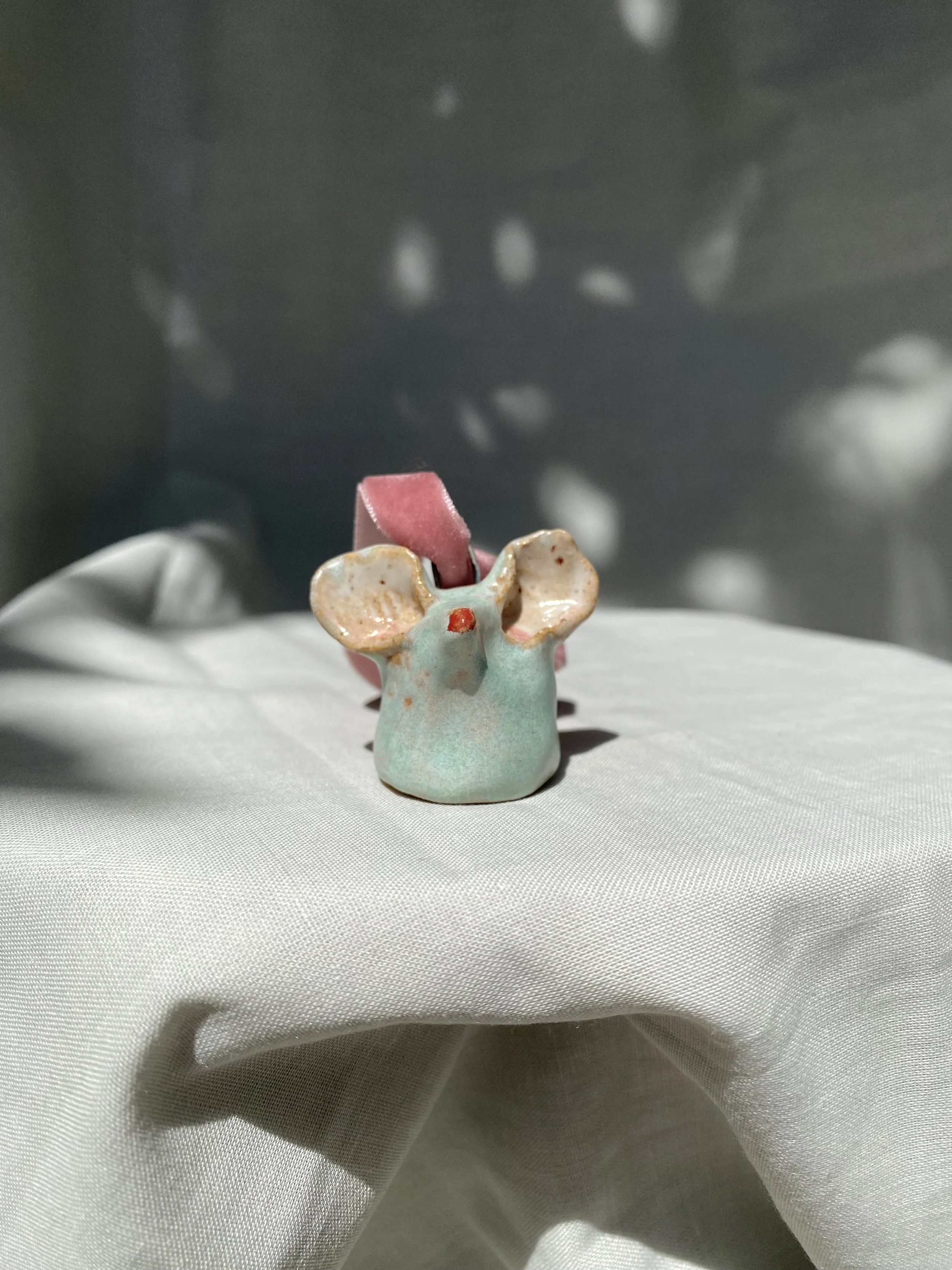 A small ceramic figurine of a light blue deer with a red nose and pink ears, placed on white fabric, with a reflective gray surface and blurred spots of light in the background.