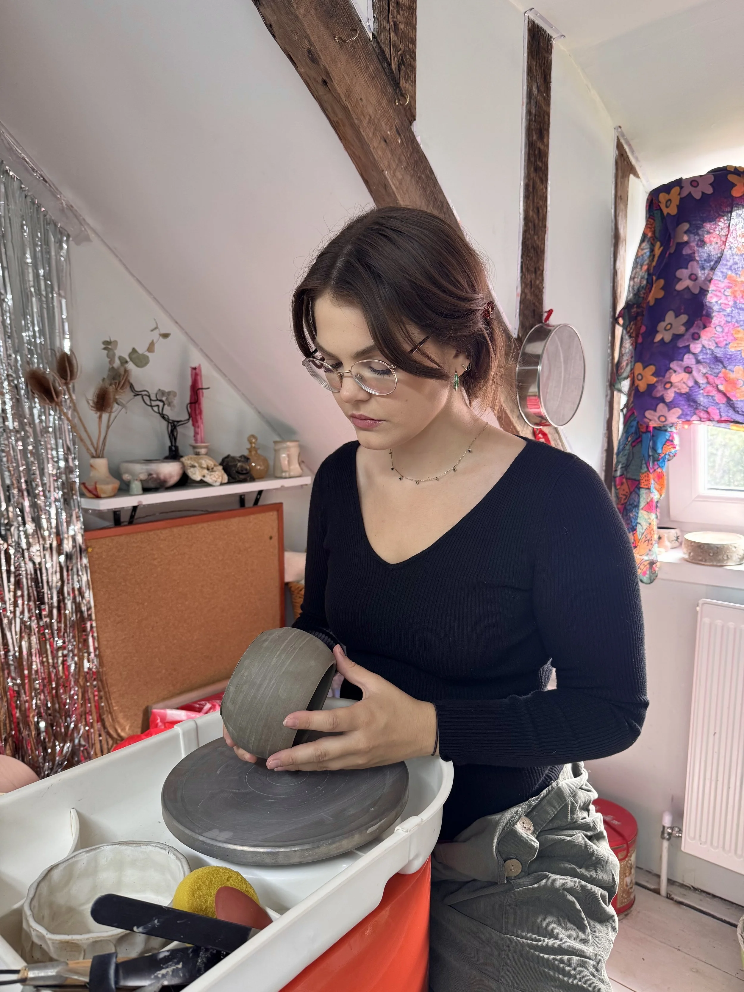 A woman with short brown hair, glasses, and a black long-sleeve shirt is sat at the pottery wheel inside a room with art supplies and decorations.