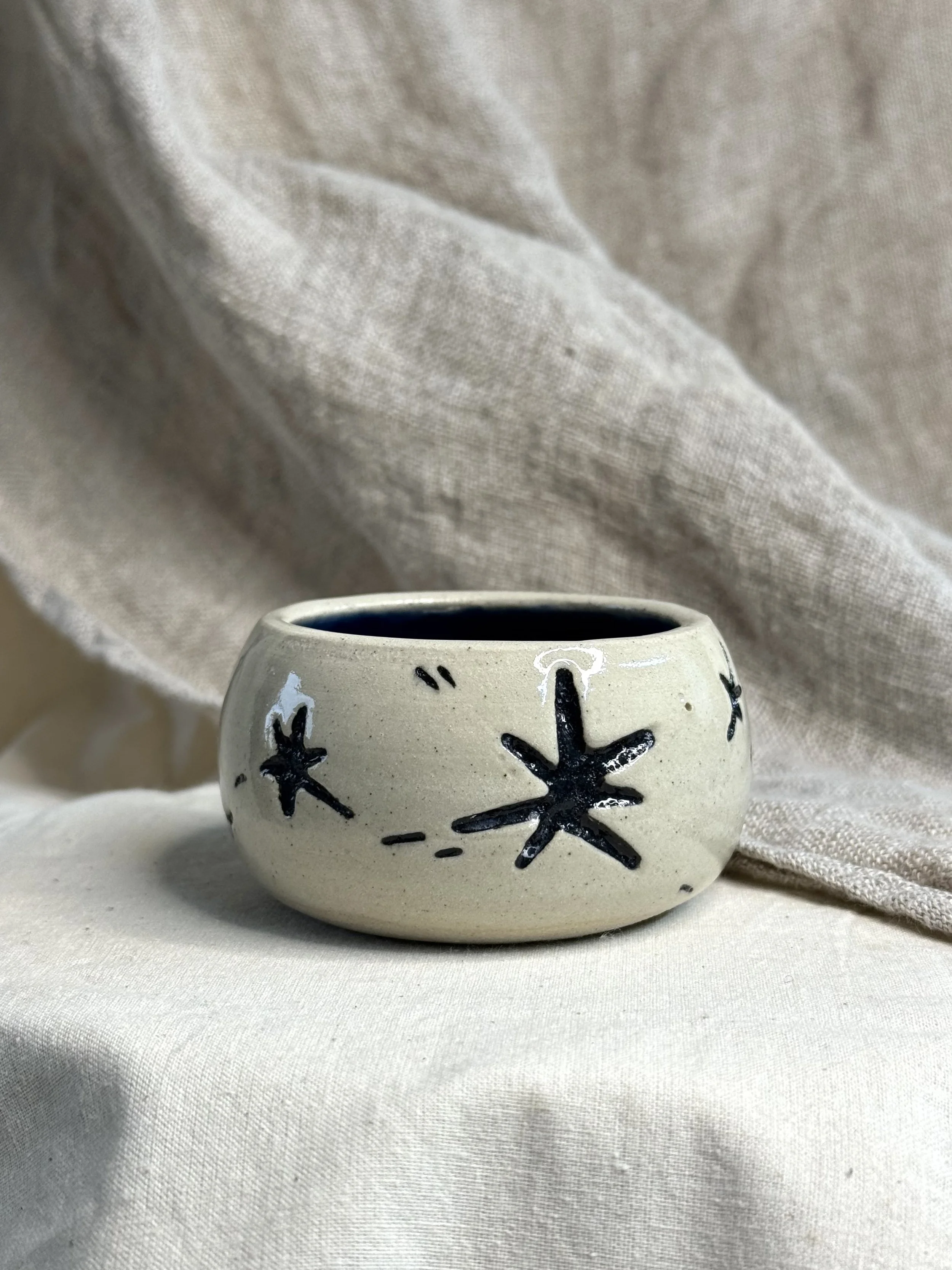 A small ceramic cup painted with black starburst designs, placed on a white fabric surface with a beige cloth background.