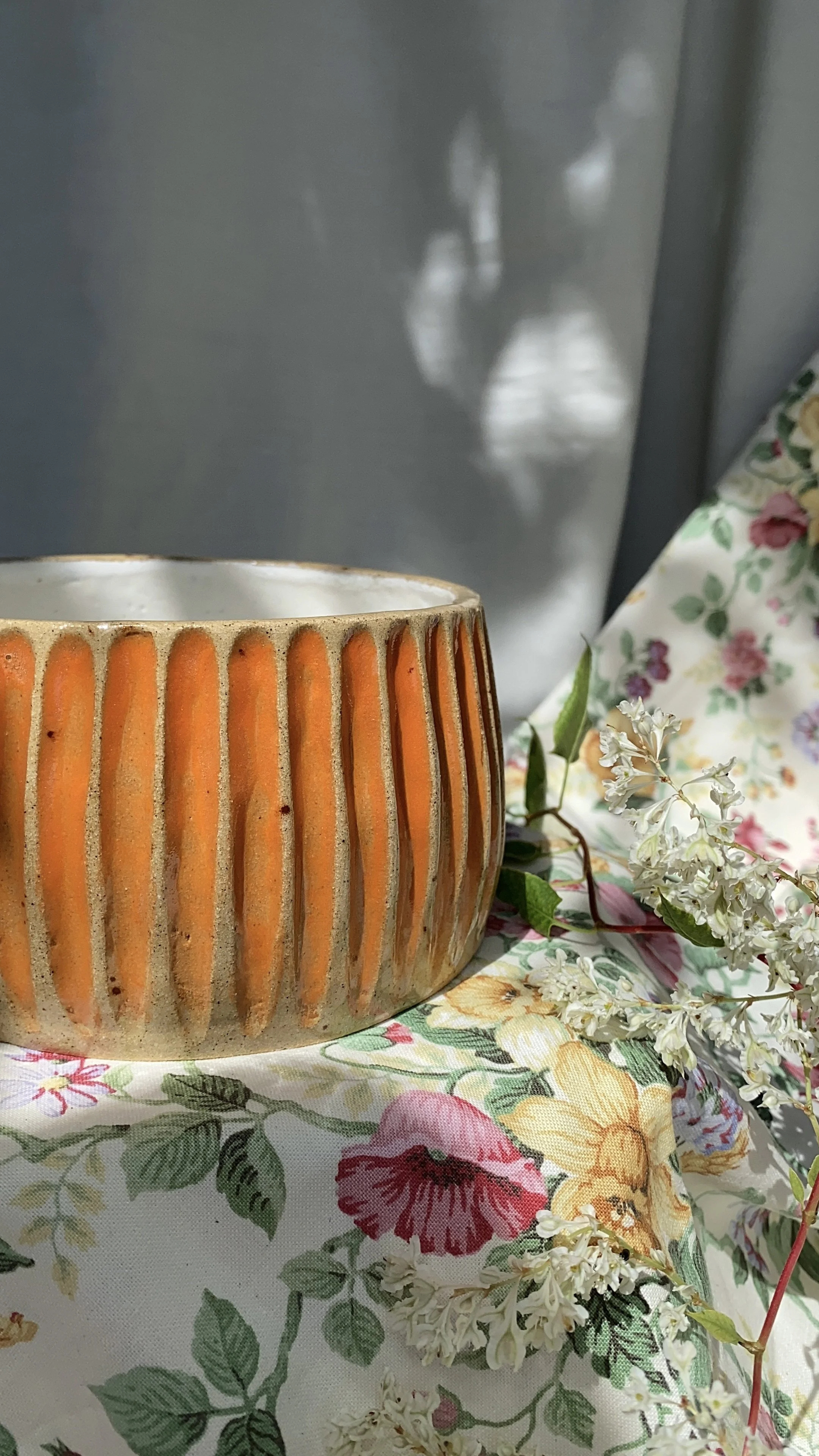 A ceramic vase with vertical orange and beige stripes on a floral cloth, accompanied by white flowers and green leaves, with a silver refrigerator in the background and a shadow of a person reflected on the fridge.