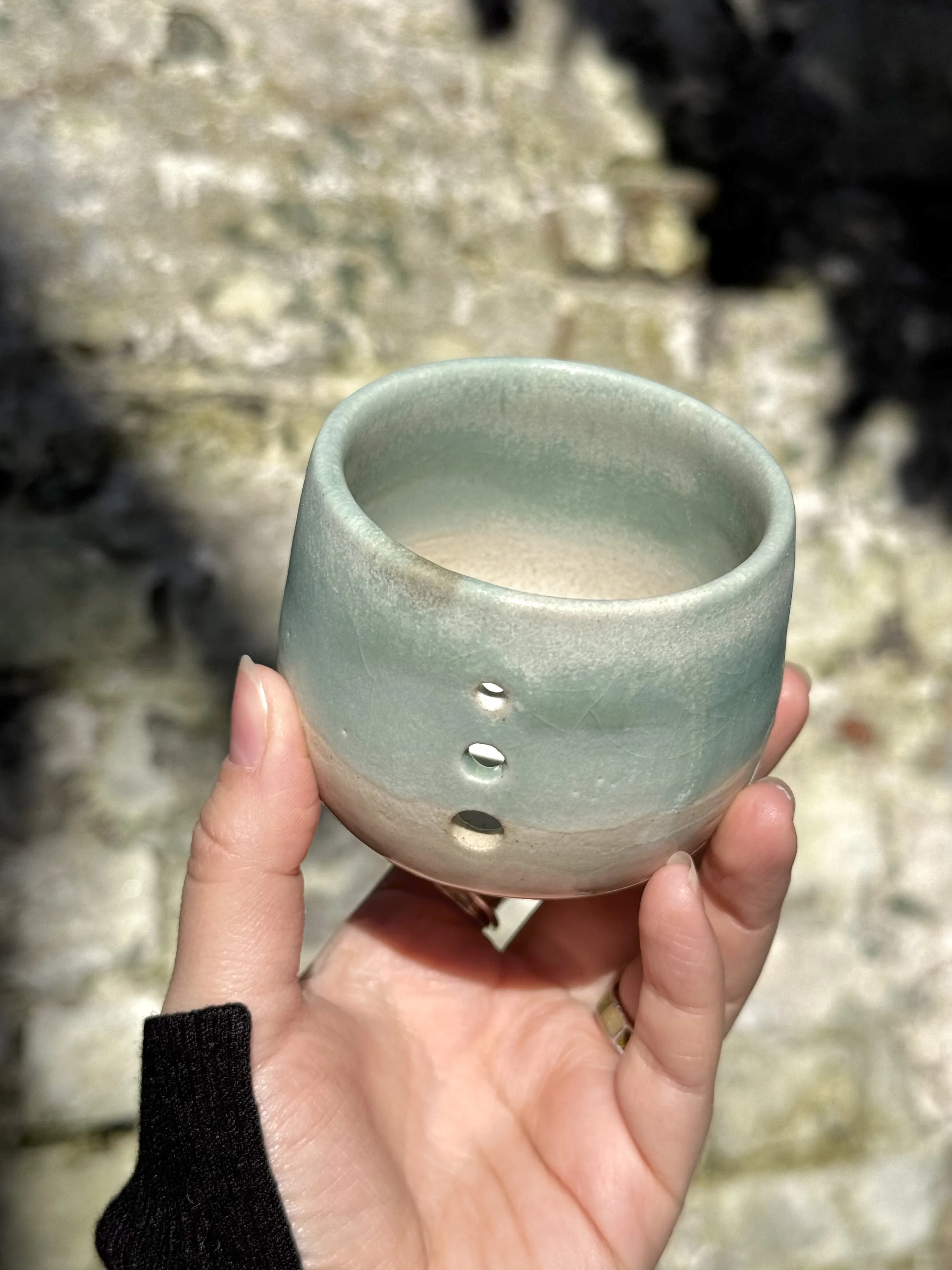 A hand holding a small ceramic cup with a rough texture and pale green glaze, with an outdoor background of rocks and moss.