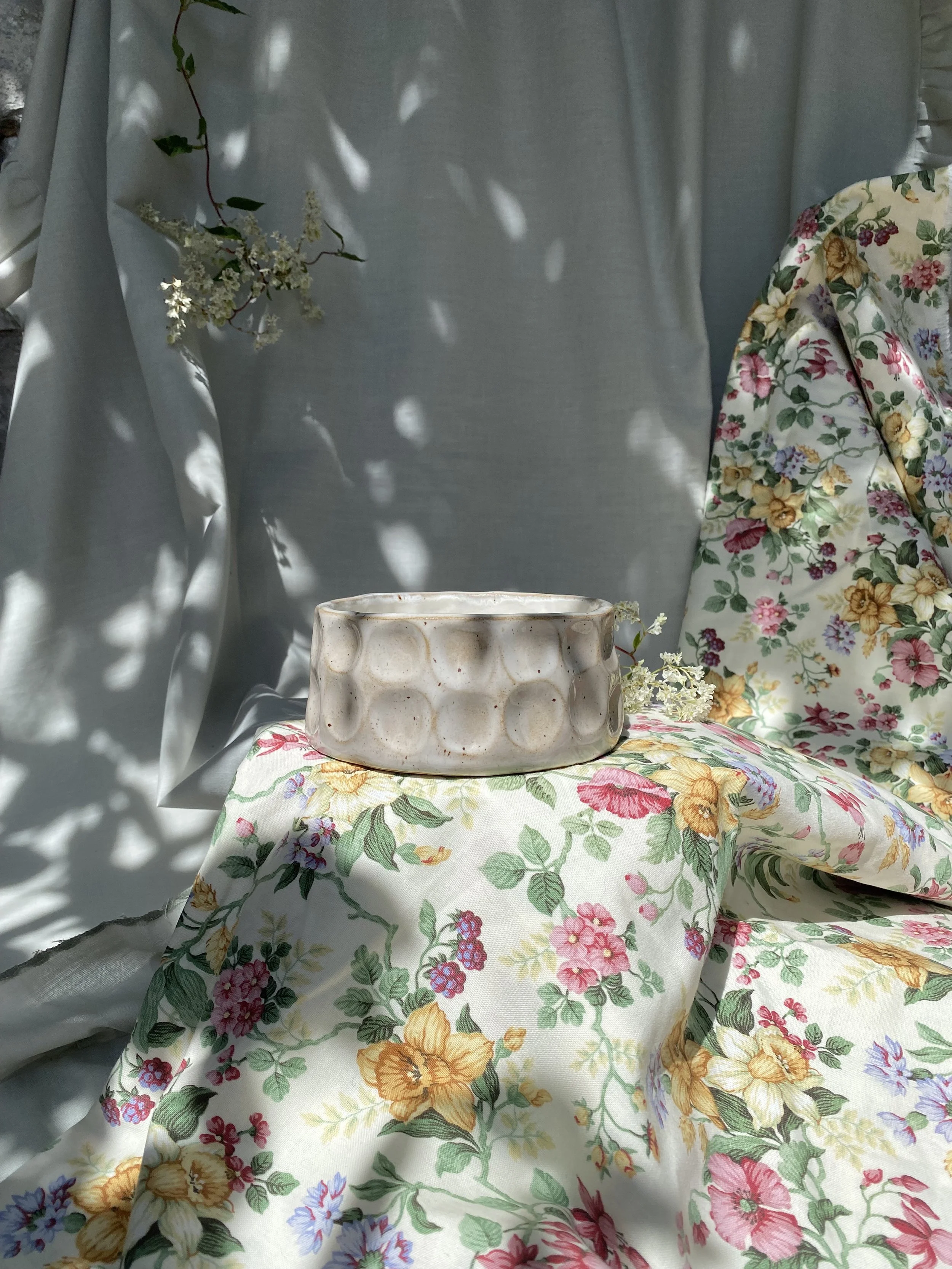 A ceramic mug with a dotted texture on a floral fabric surface, with white flowers and green leaves, and a light gray backdrop with shadows of leaves.