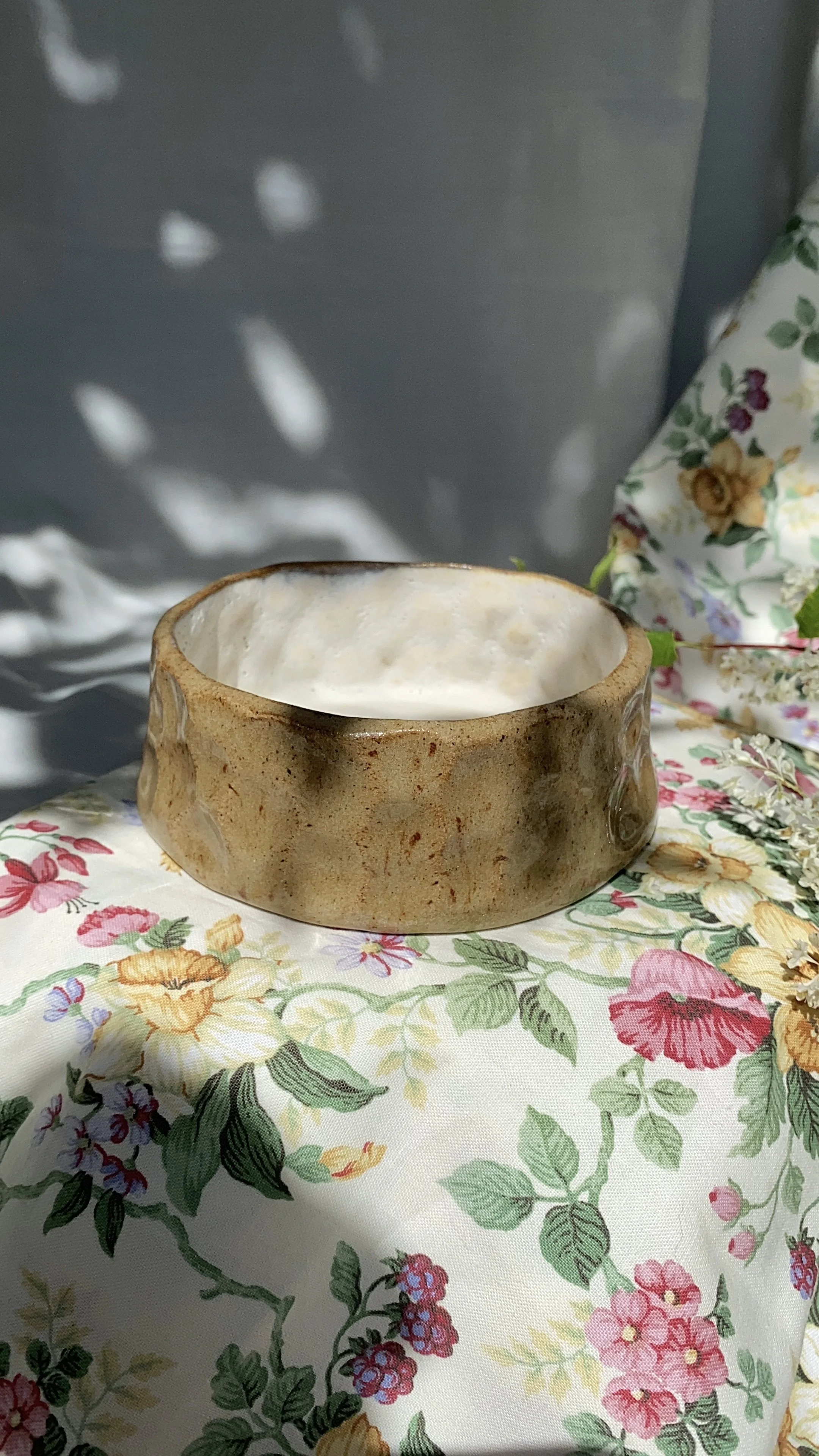 A round, rustic, brown ceramic bowl with a glossy white interior, placed on a floral-patterned fabric with colorful flowers and green leaves. In the background, there is a gray metallic surface and part of a floral-patterned cloth.