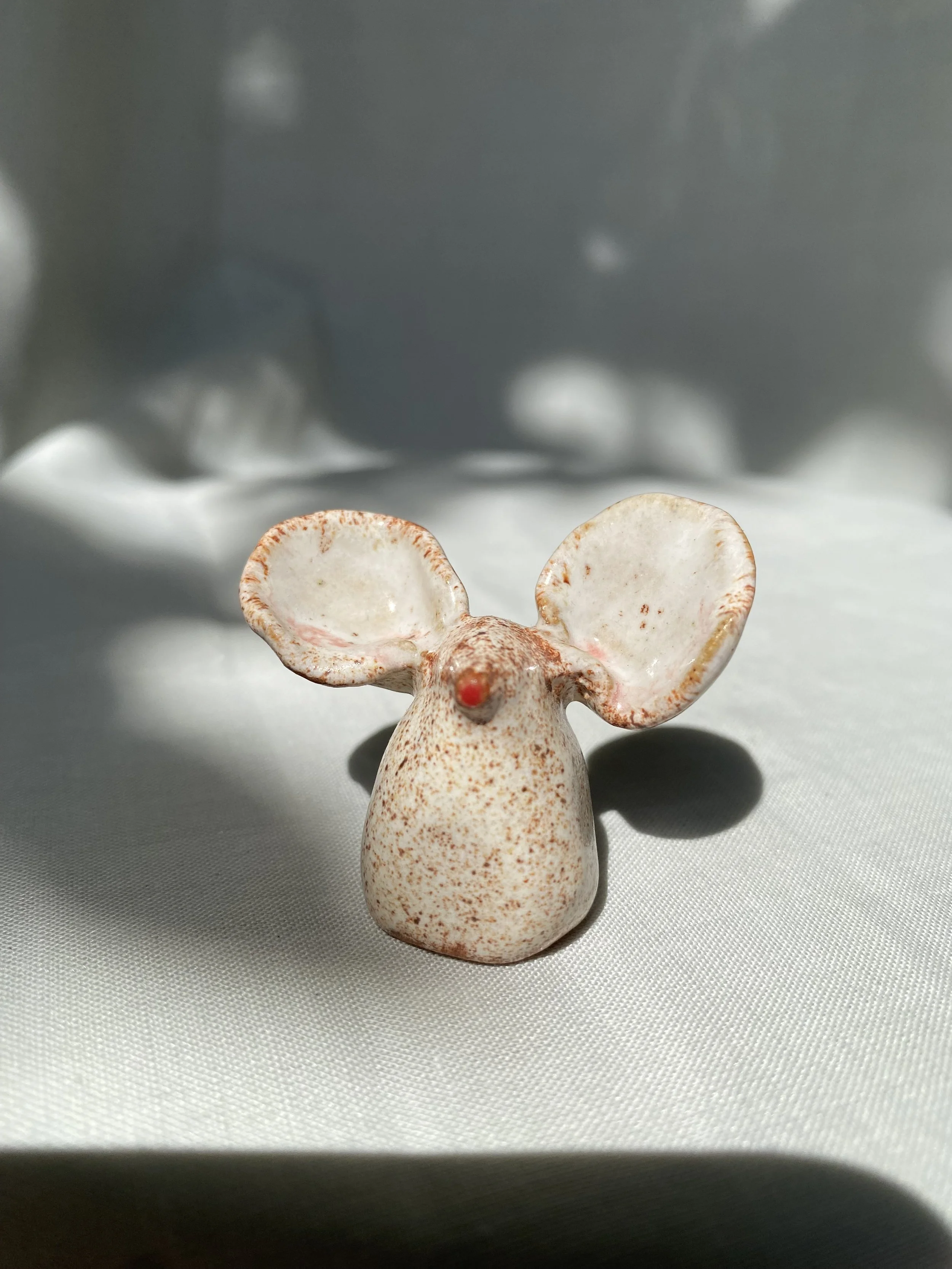 Small decorative figurine of an animal, possibly a mouse, with large ears, a red nose, and speckled pinkish-white body.