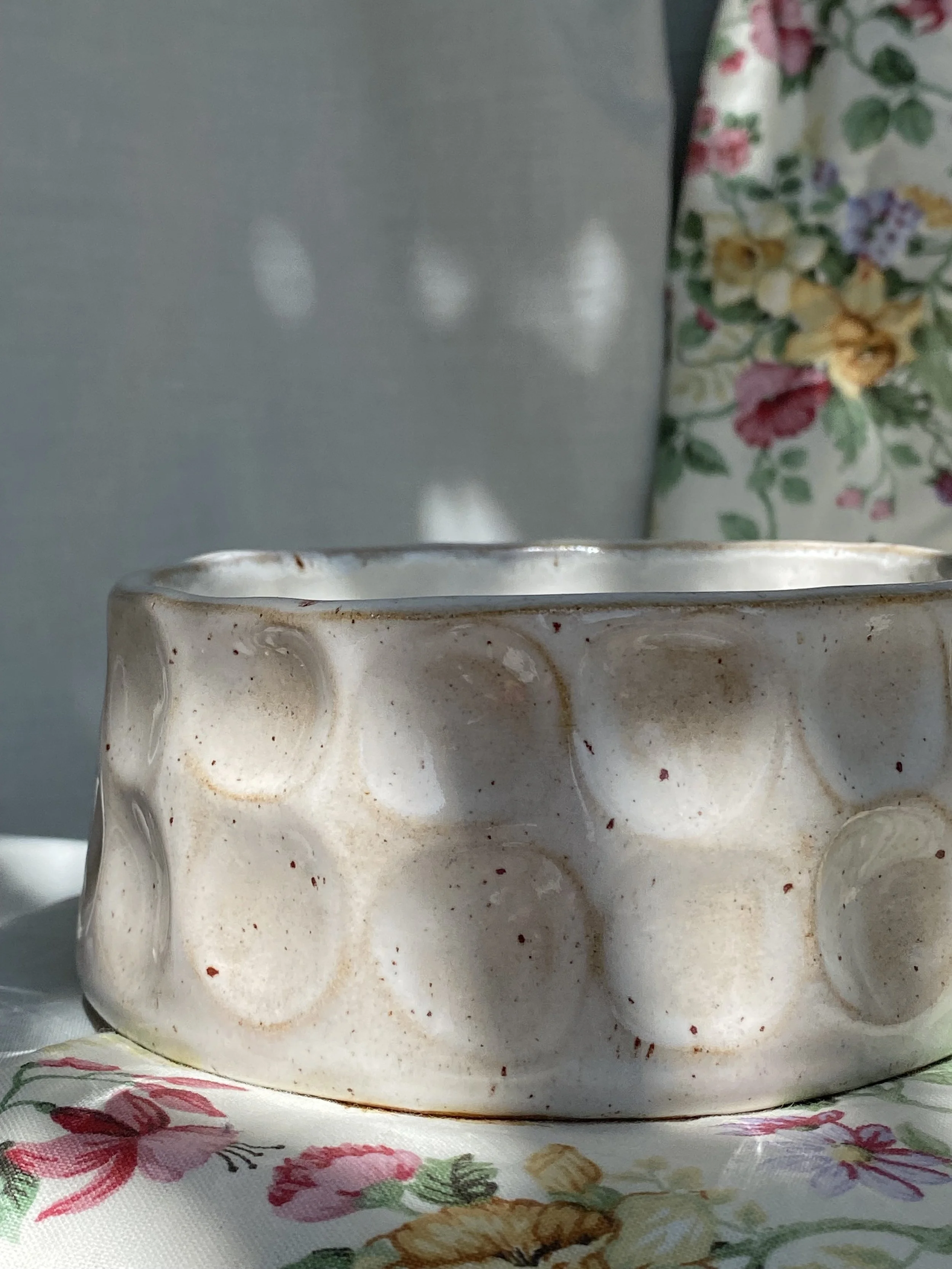 Close-up of a ceramic bowl with a white and brown speckled glaze sitting on a floral tablecloth.