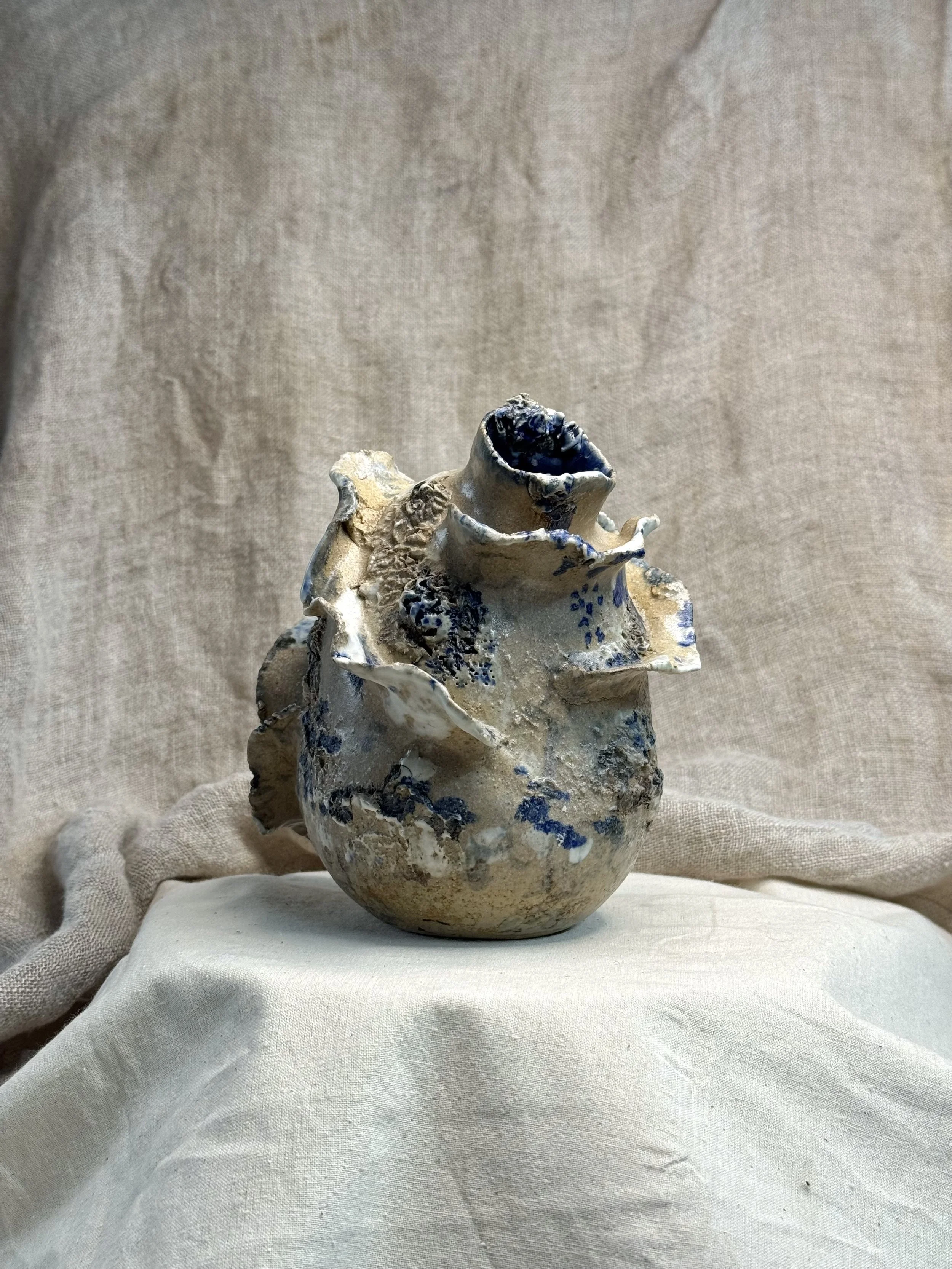 A ceramic vase with irregular, textured edges and a rough surface, decorated with blue and white glaze, placed on a light fabric background.