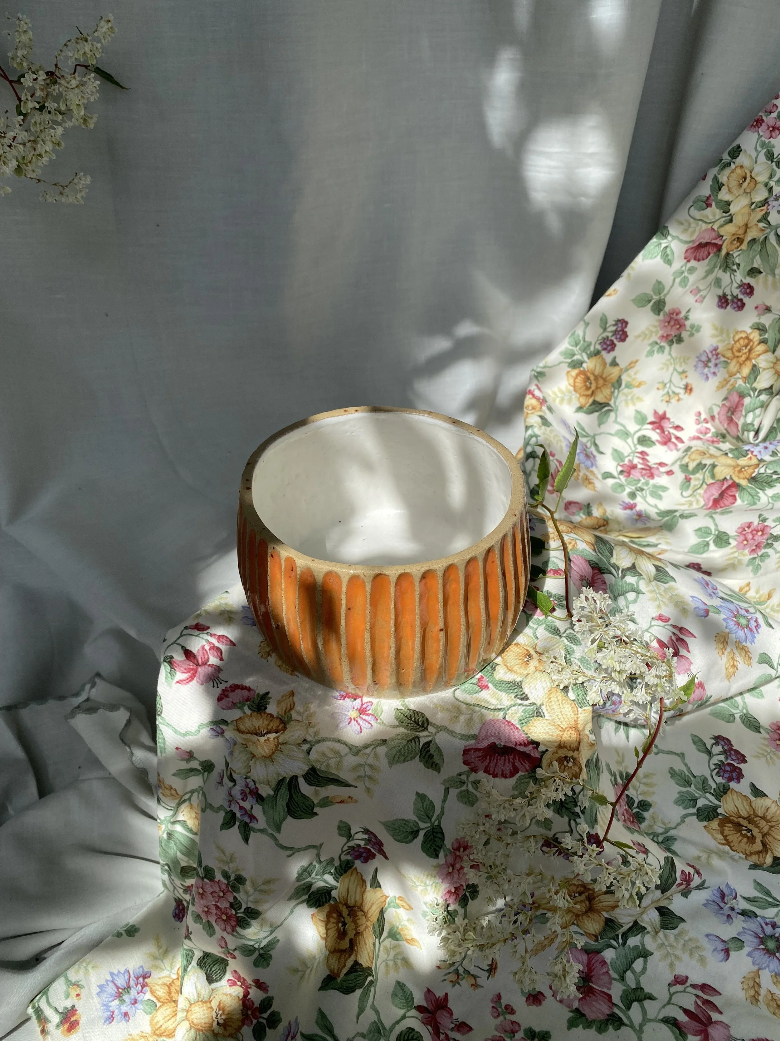 A ceramic cup with orange vertical stripes on a floral fabric with white, pink, yellow, purple, and green patterns. The fabric is draped over a surface, and a sprig of white flowers is placed next to the cup. The background consists of a light-colore