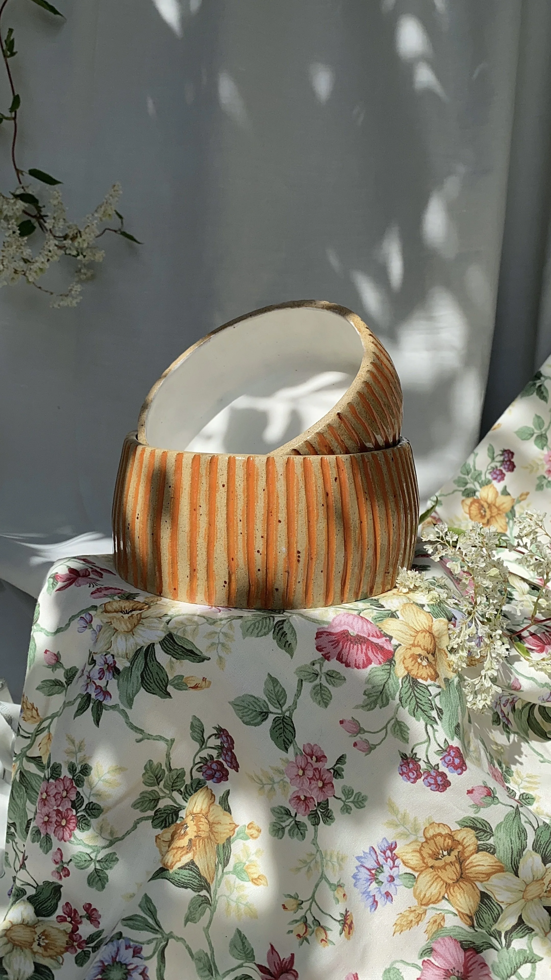 Two ceramic bowls with orange and beige vertical stripes stacked on a floral fabric tablecloth, surrounded by white flowers, with soft sunlight and shadows in the background.