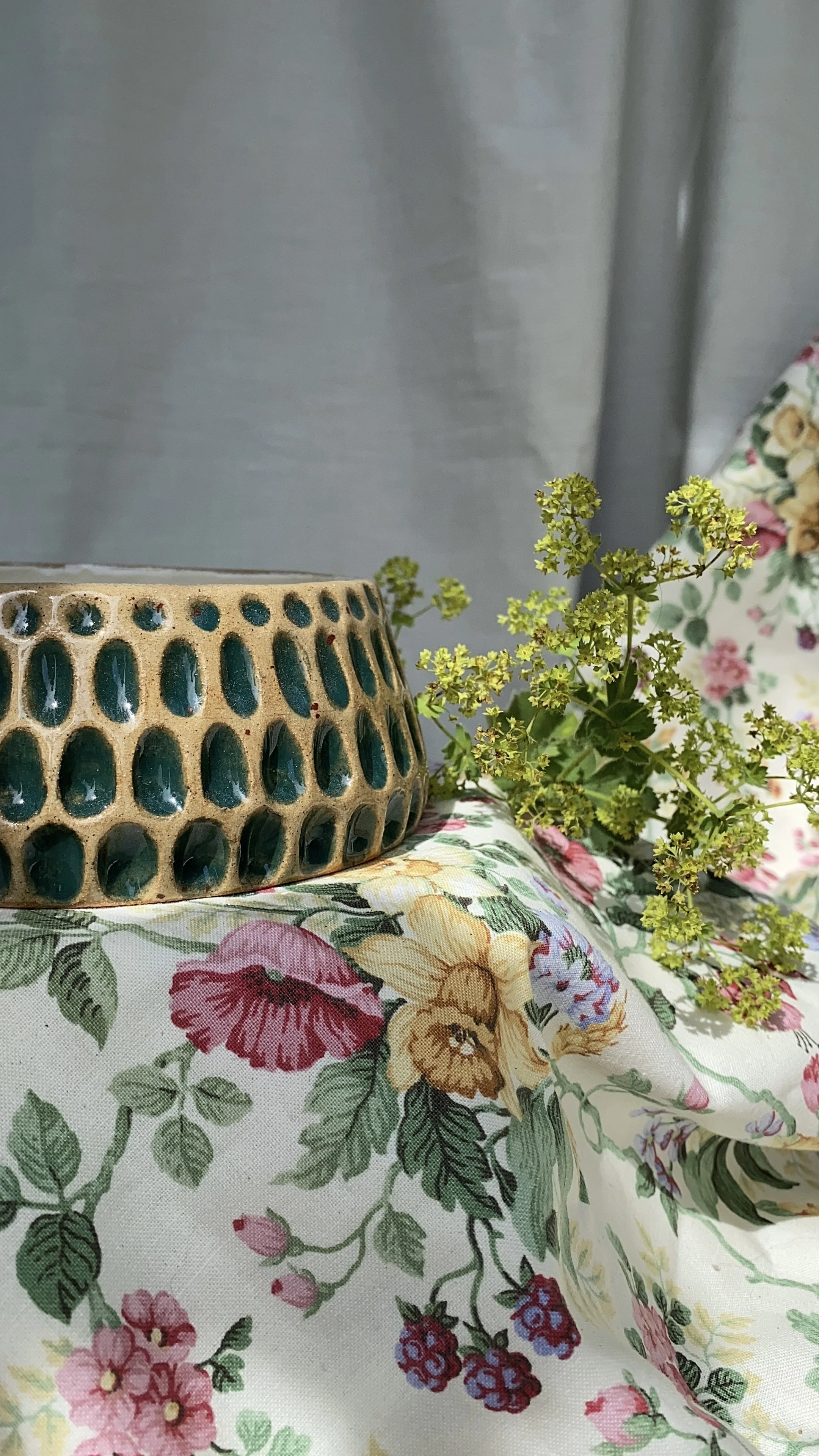 Ceramic bowl with turquoise interior holes, floral tablecloth with pink, yellow, and green pattern, some green foliage, and sheer gray curtain in background.