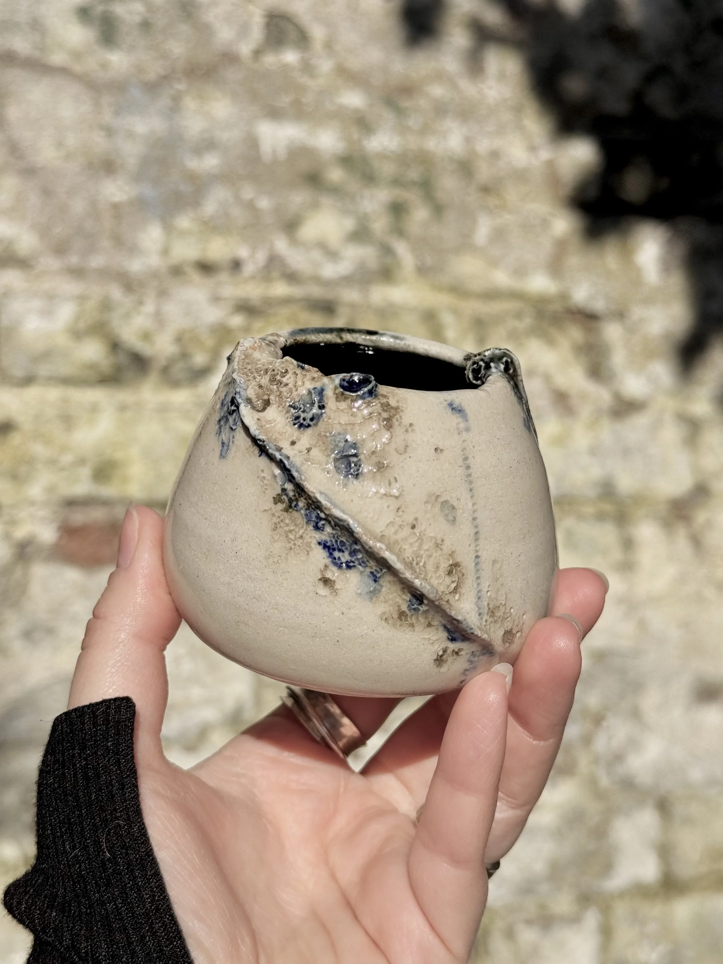 A hand holding a beige ceramic cup with a dark interior and a decorative black line that appears to be melting or dripping down the side, against a blurred rocky background.