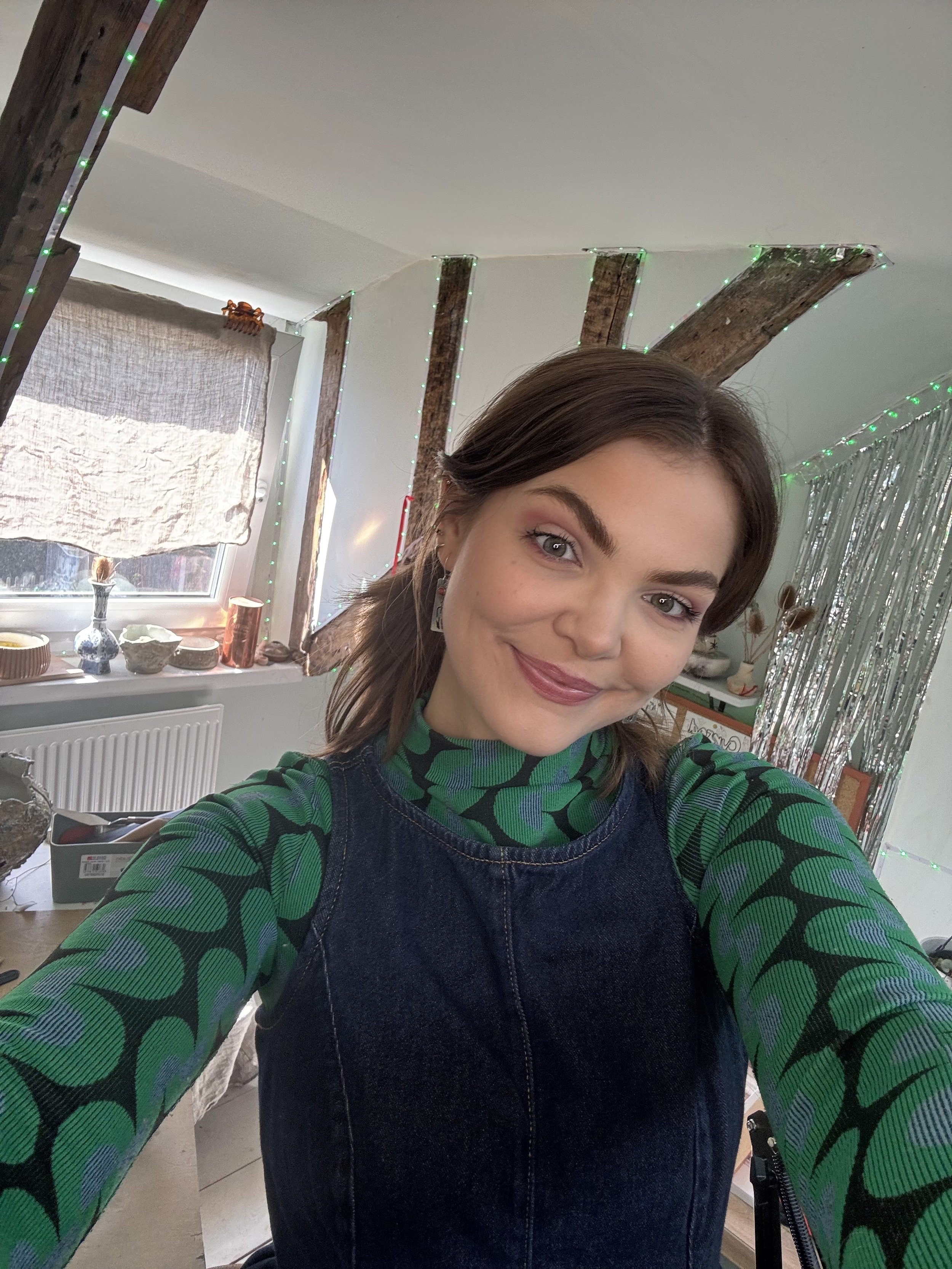 Young woman taking a selfie in a decorated room with wooden beams, LED string lights, and a window with a curtain.