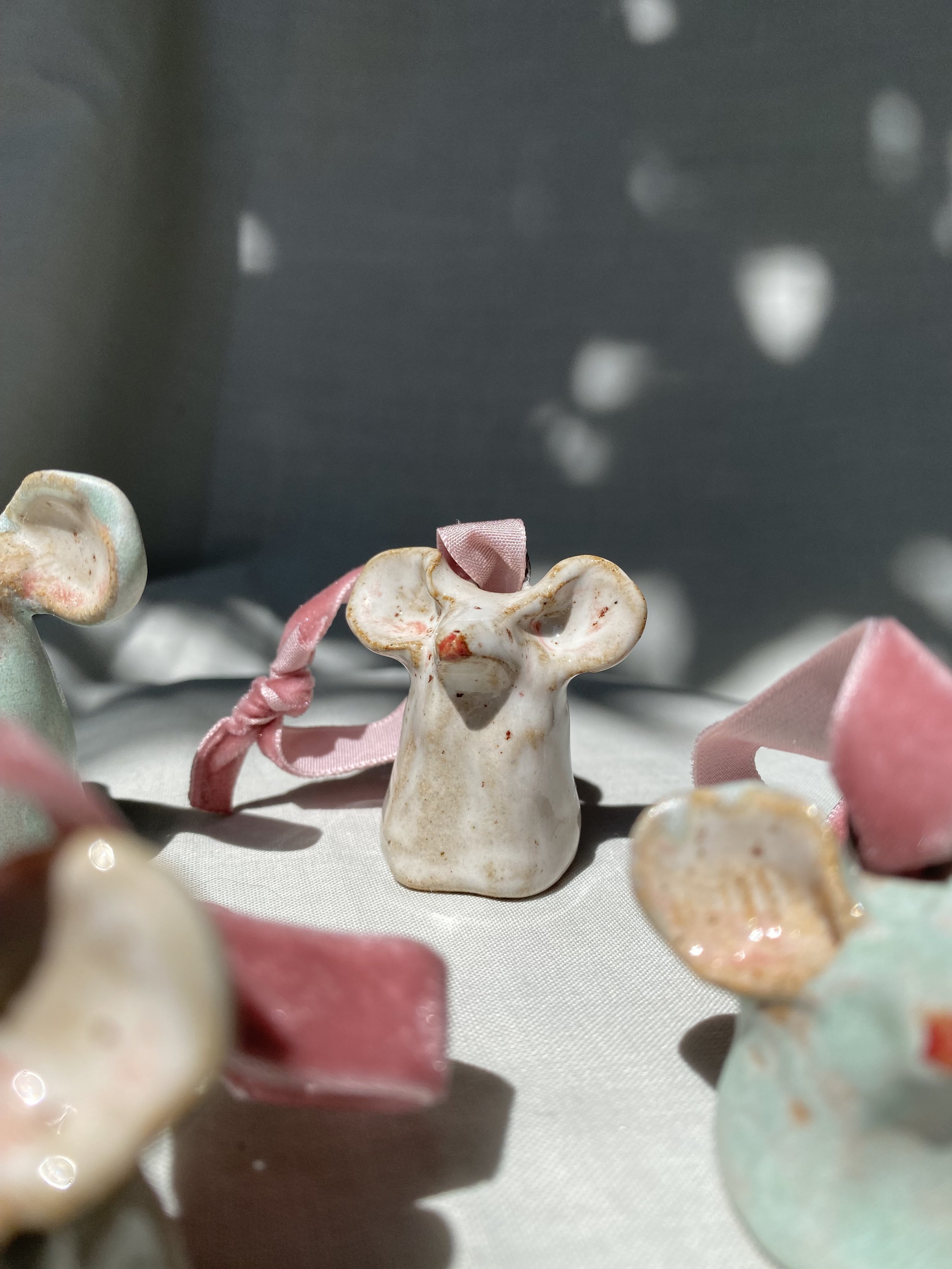 Close-up of a small ceramic angel figurine with a pink ribbon, surrounded by other small ceramic decorations, with a blurred background.