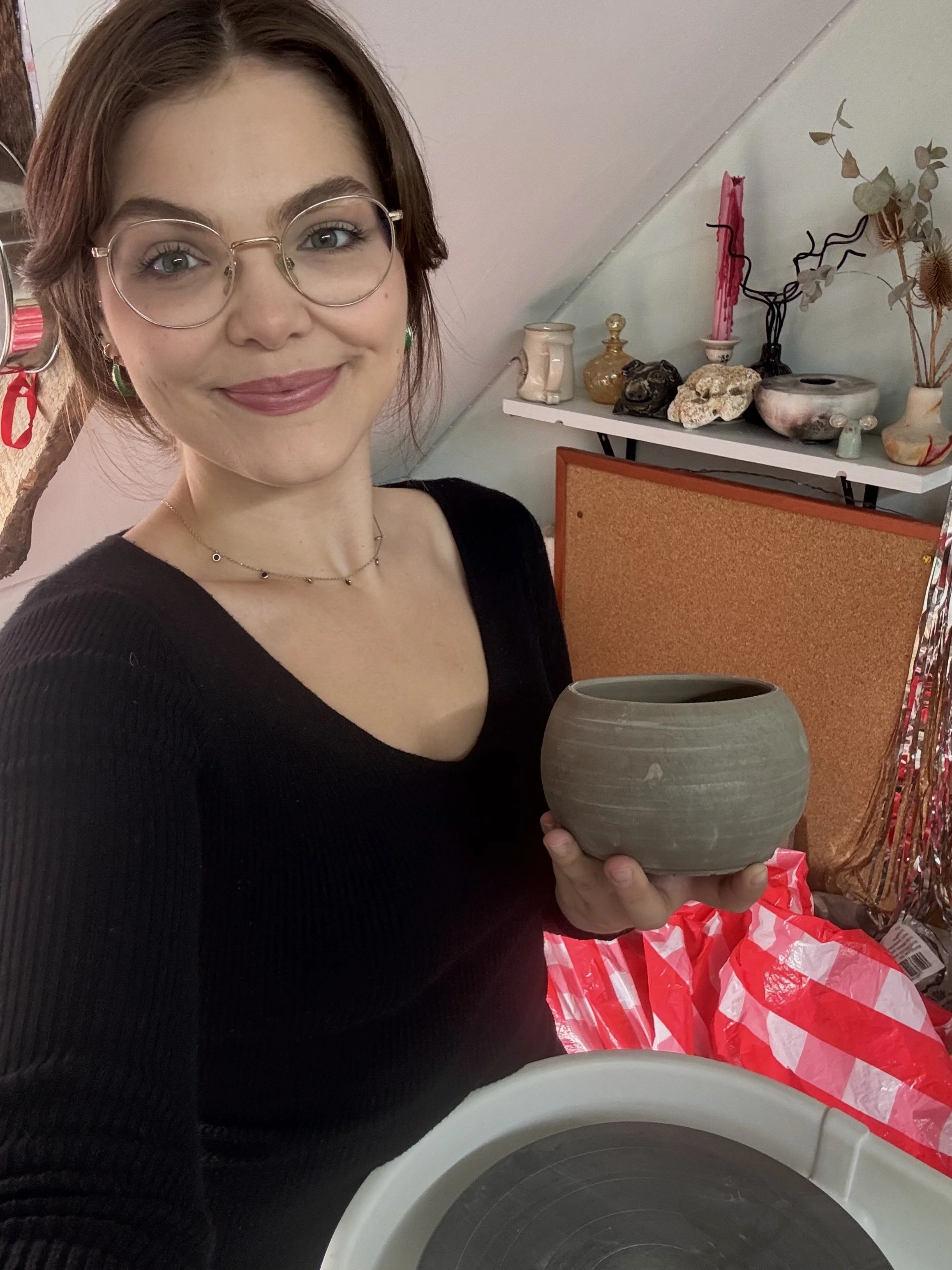 A woman with glasses and wearing a black shirt holding a earthenware pot, standing in a room with decorative items and red and white checkered bags in the background.