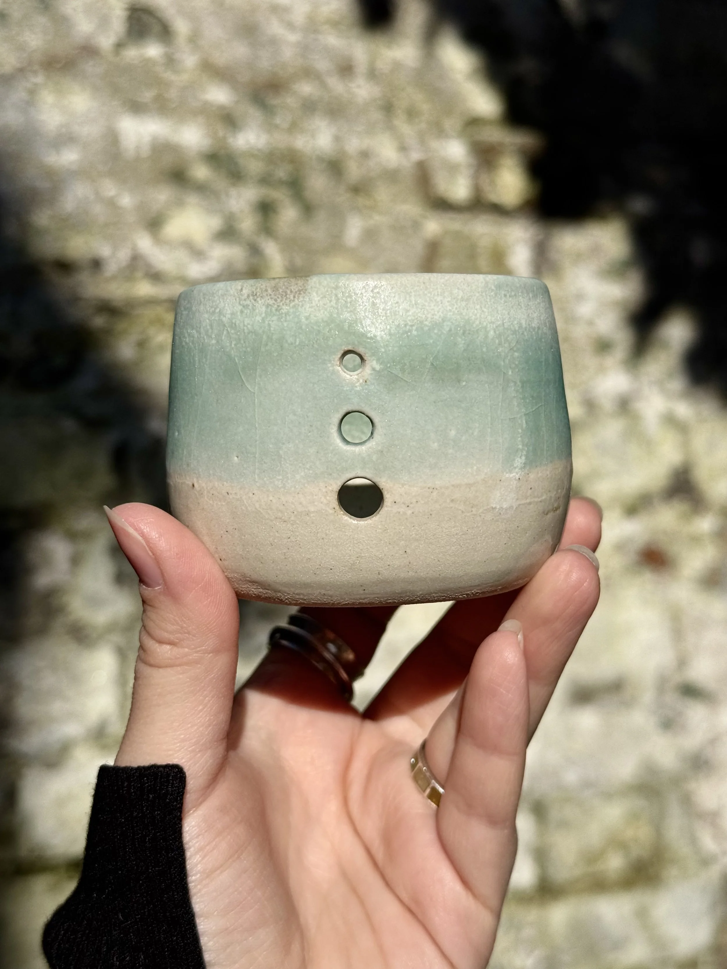 A person's hand holding a small, light-colored ceramic bowl with four circular holes on its side, against a blurred outdoor background.