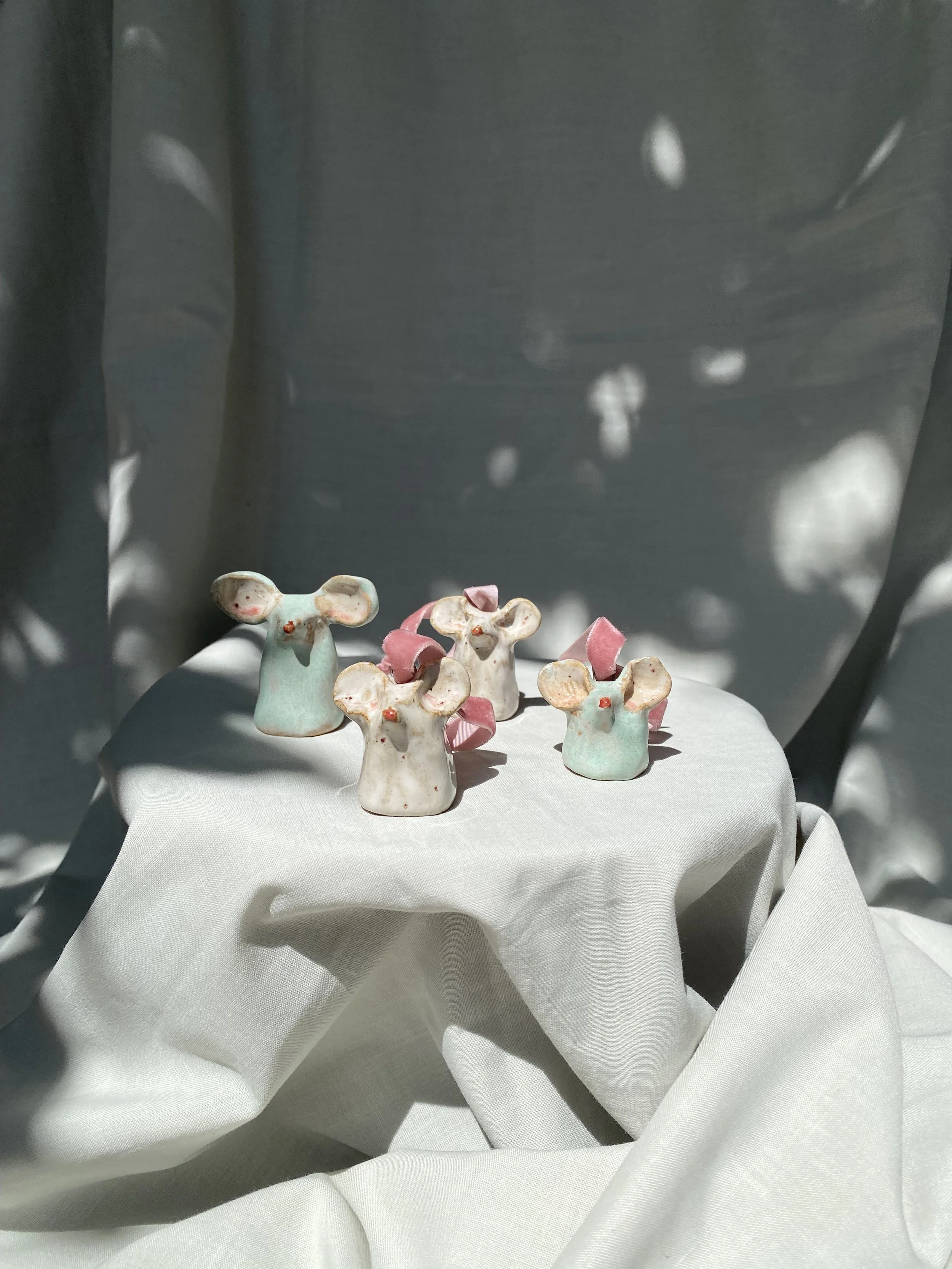 Ceramic figurines of mice with pink and blue accents displayed on a white fabric-covered surface, with a reflective metallic background creating light spots.