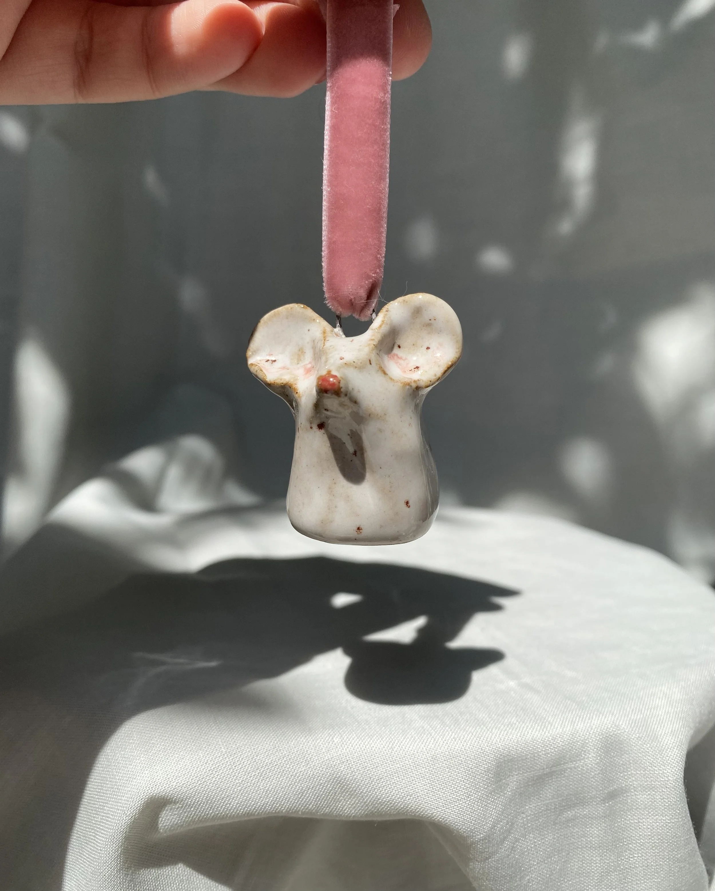 A small, white ceramic vessel shaped like a bear, supported by a pink, soft, fuzzy stick held by a person's fingers, with a neutral background and the vessel casting a shadow on a white surface.