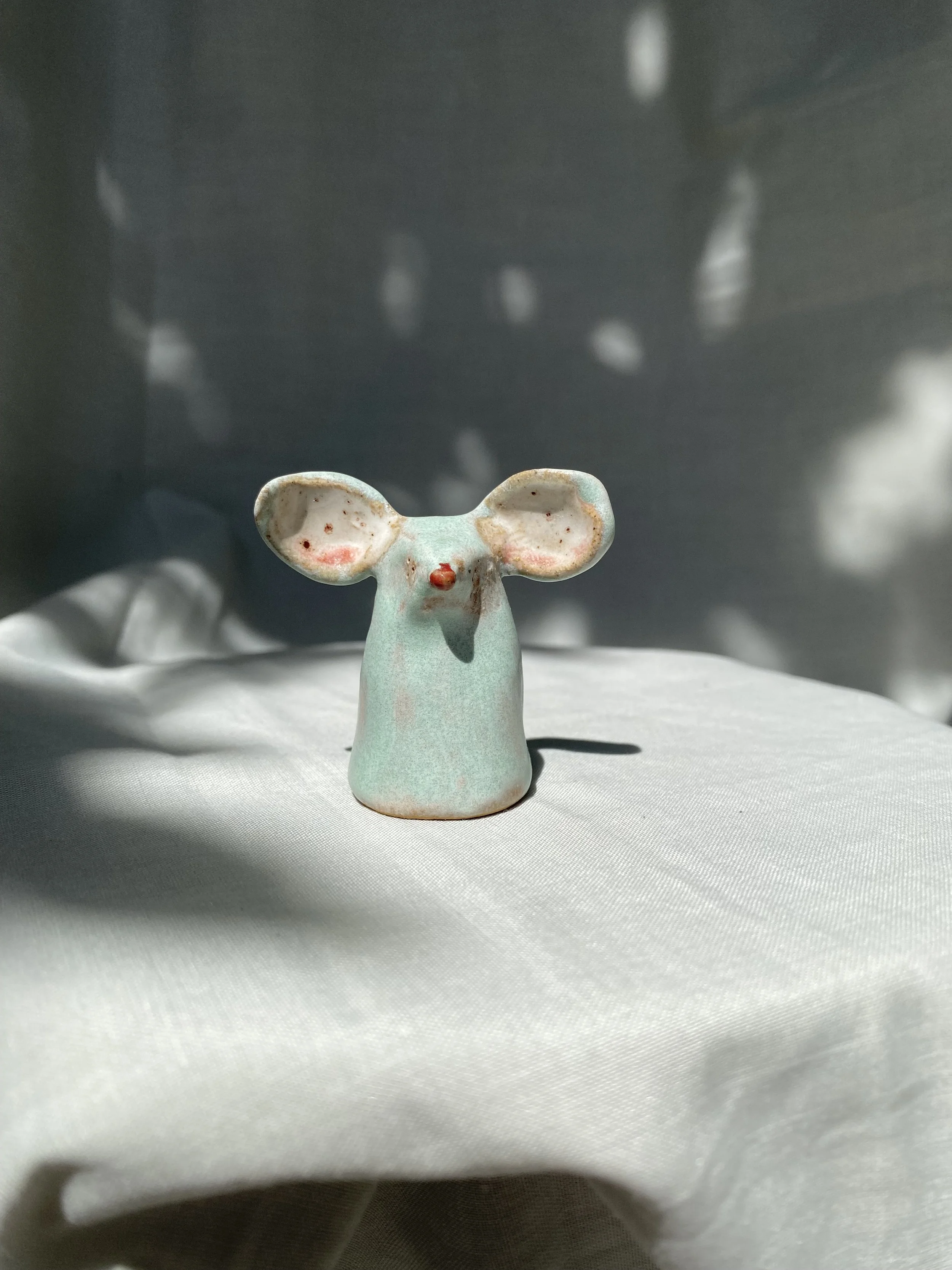 Small ceramic figurine of a mouse with pink cheeks, standing on a white fabric surface, dark background with blurred lights.