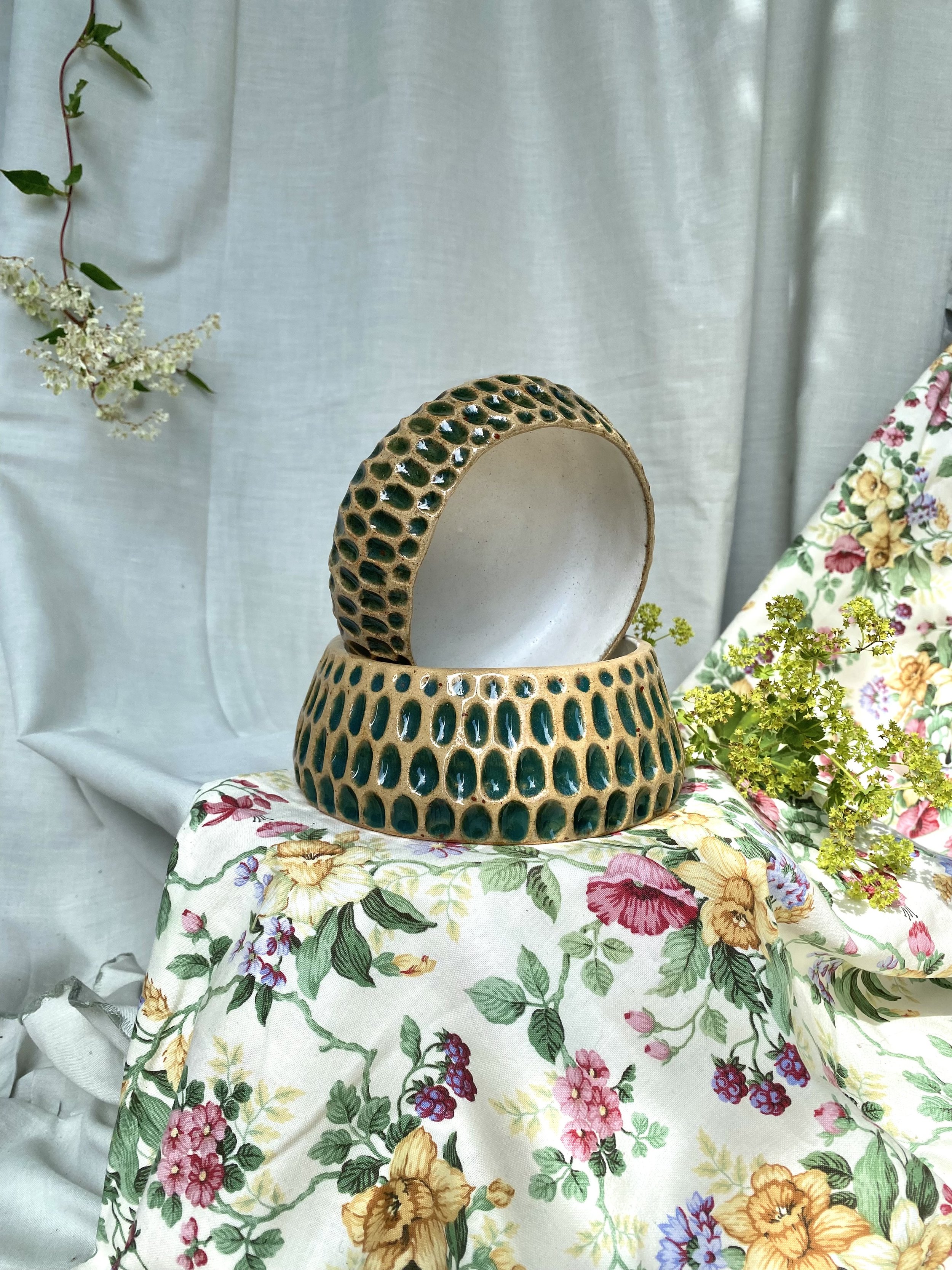 A ceramic vase with a narrow neck and decorative holes, placed on a floral fabric with plants and a floral patterned backdrop.