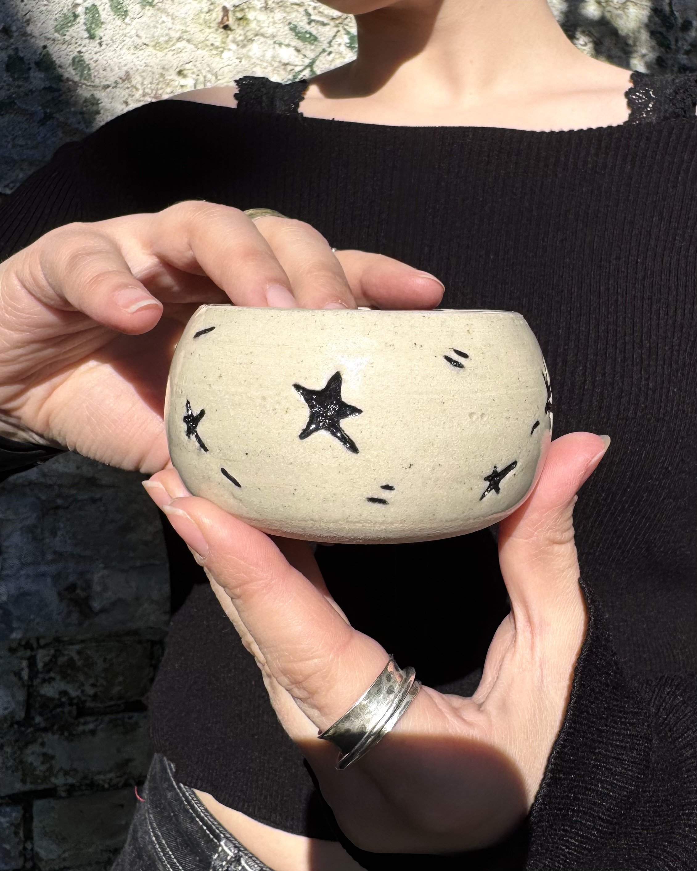 A small ceramic carved vase decorated with black star designs. The person is wearing a black ribbed top with lace shoulder details and patterned pants, standing outdoors with a textured stone background.