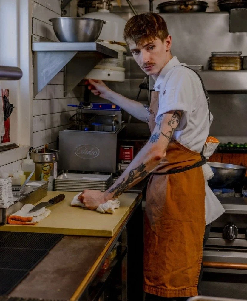 A male chef with tattoos on his arms, wearing a white shirt and orange apron, is preparing food in a kitchen.