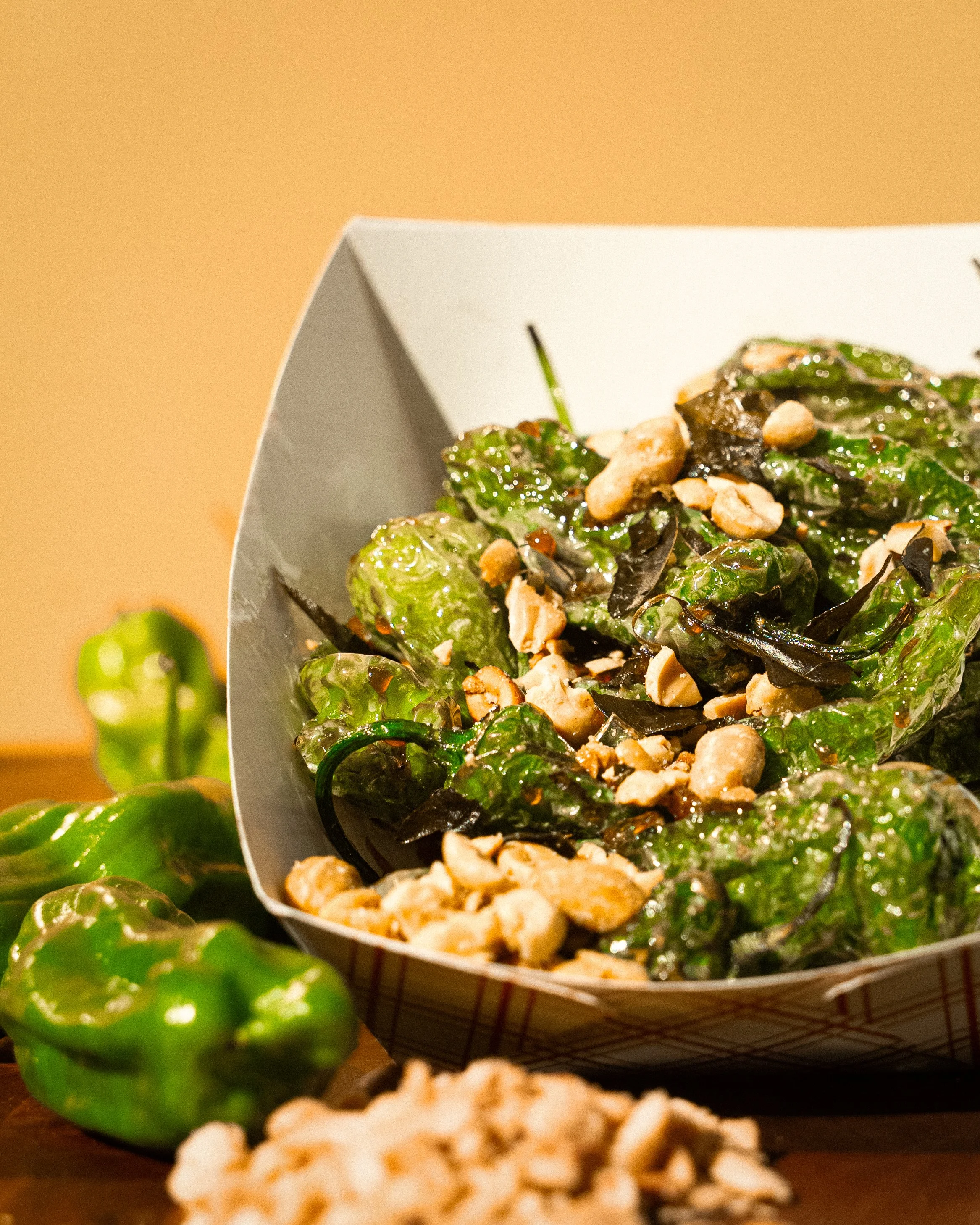 Close-up of a salad with greens, nuts, and possibly dried fruit in a paper tray, with green Bell peppers in the foreground and a blurred brown background.