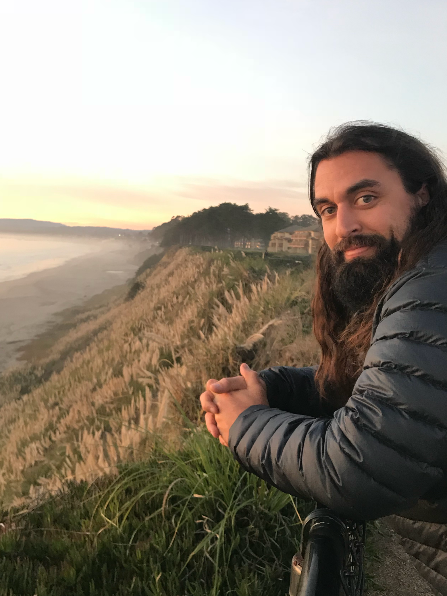 A man with long dark hair and a beard kneeling on a grassy area near the coast, with houses on a hill in the background during sunset.