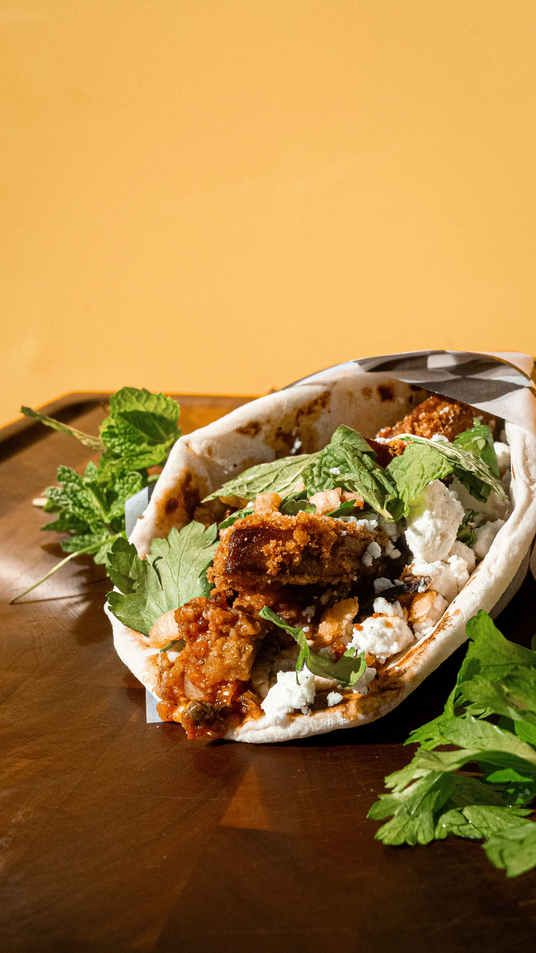 A close-up of a taco filled with fried chicken, cheese, and herbs on a wooden tray.