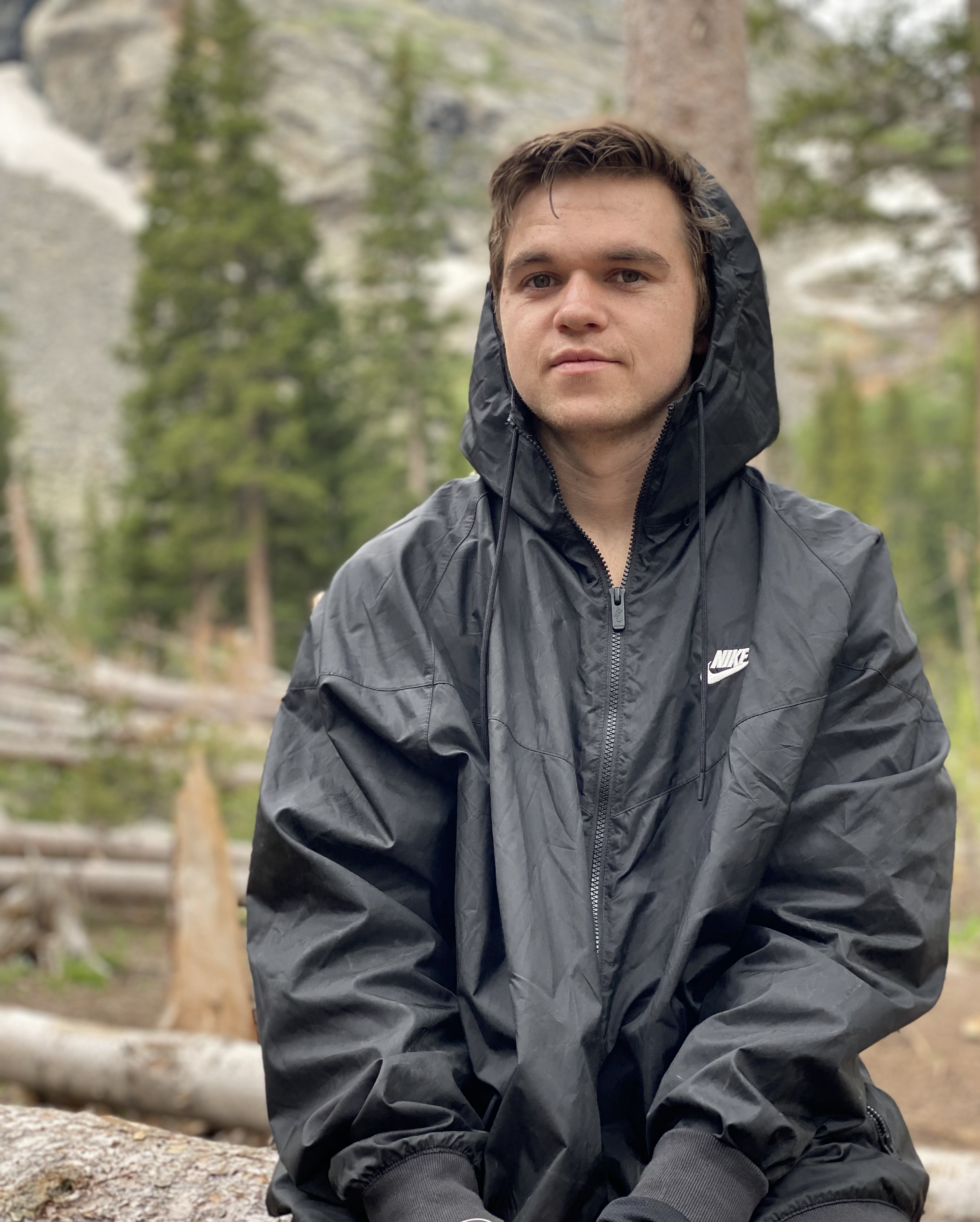 A young man with short brown hair, wearing a black Nike windbreaker jacket with the hood up, sitting outdoors in a forested area with trees and rocks in the background.