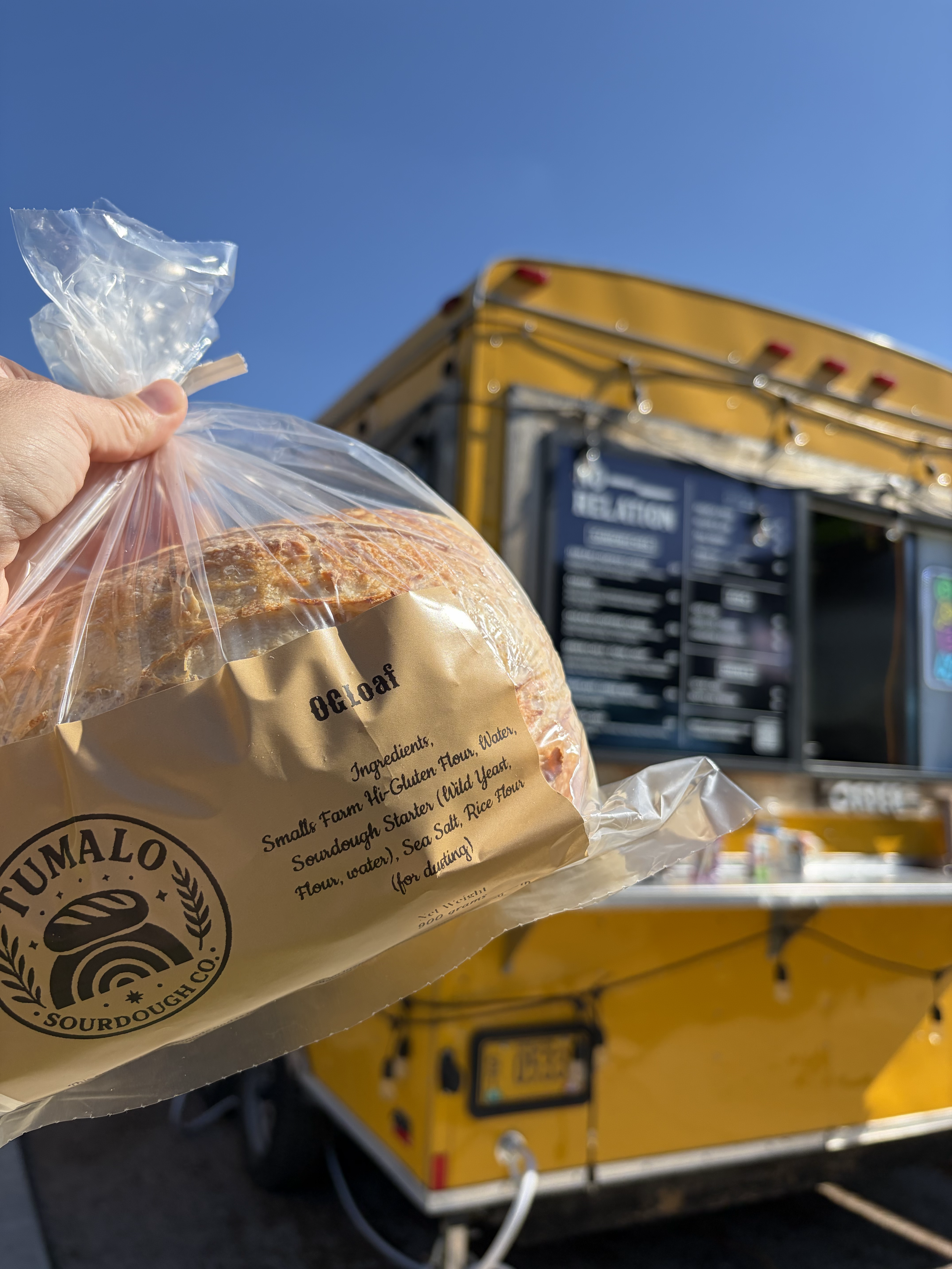 A hand holding a plastic bag with a loaf of bread in front of a yellow food truck with a black menu board.