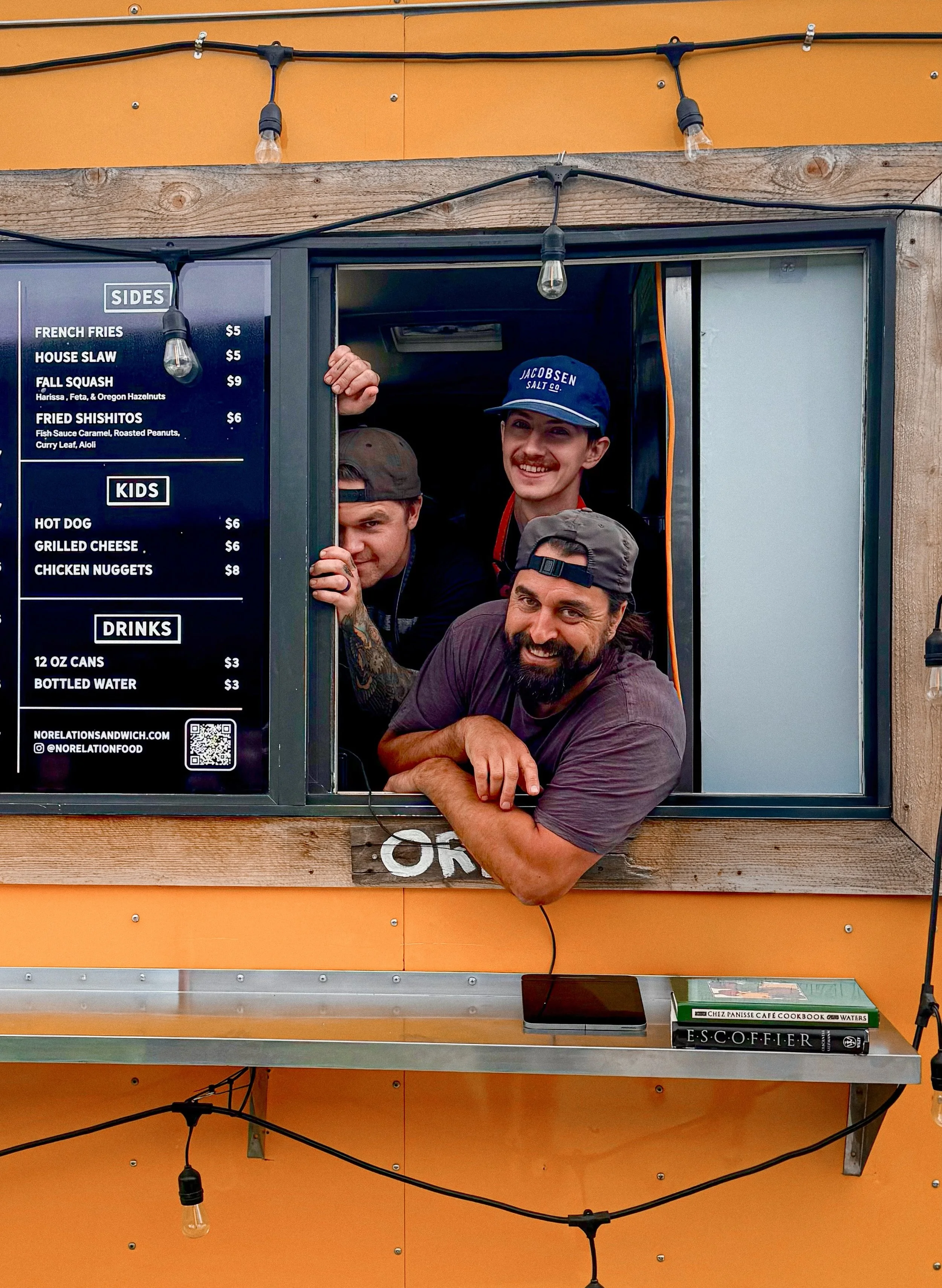 Three men leaning out of a food truck window, smiling. One wears a blue cap reading 'JACOBSEN SALT CO.', another has a gray hat, and the third has a backward gray cap. The food truck has a black menu listing sides, kids' meals, and drinks, with orange and wooden elements in the background.