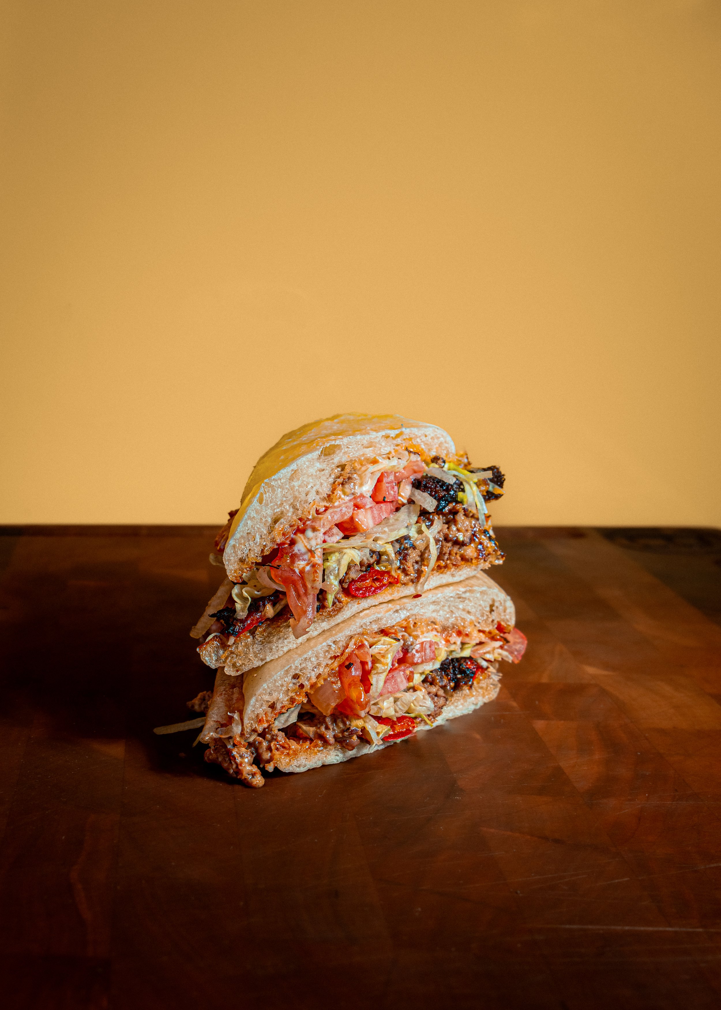 A sandwich cut in half and stacked on a wooden cutting board, filled with meat, leafy greens, and cheese.