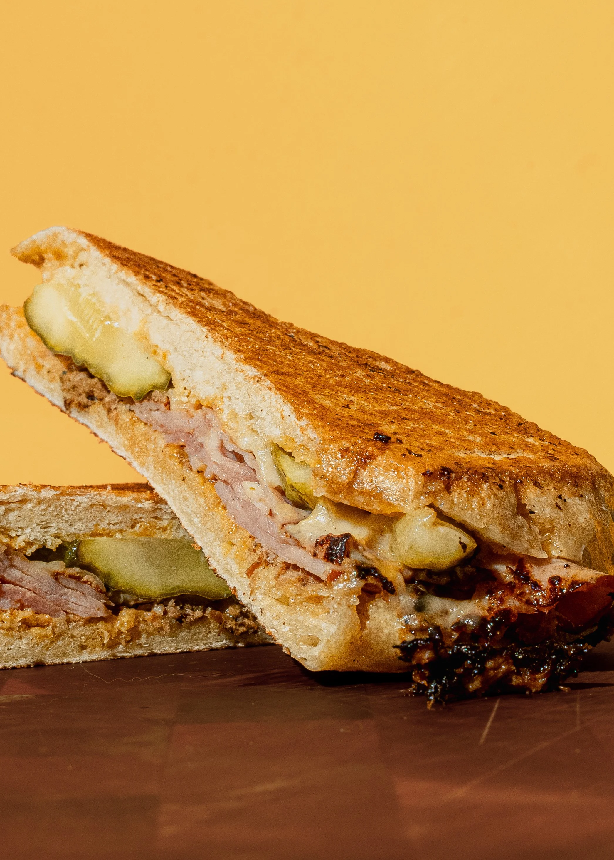 Close-up of a toasted sandwich with melted cheese, ham, and pickles in a toasted bread triangle, with sauce and melted cheese spilling out at the bottom, on a dark wooden surface against a yellow background.