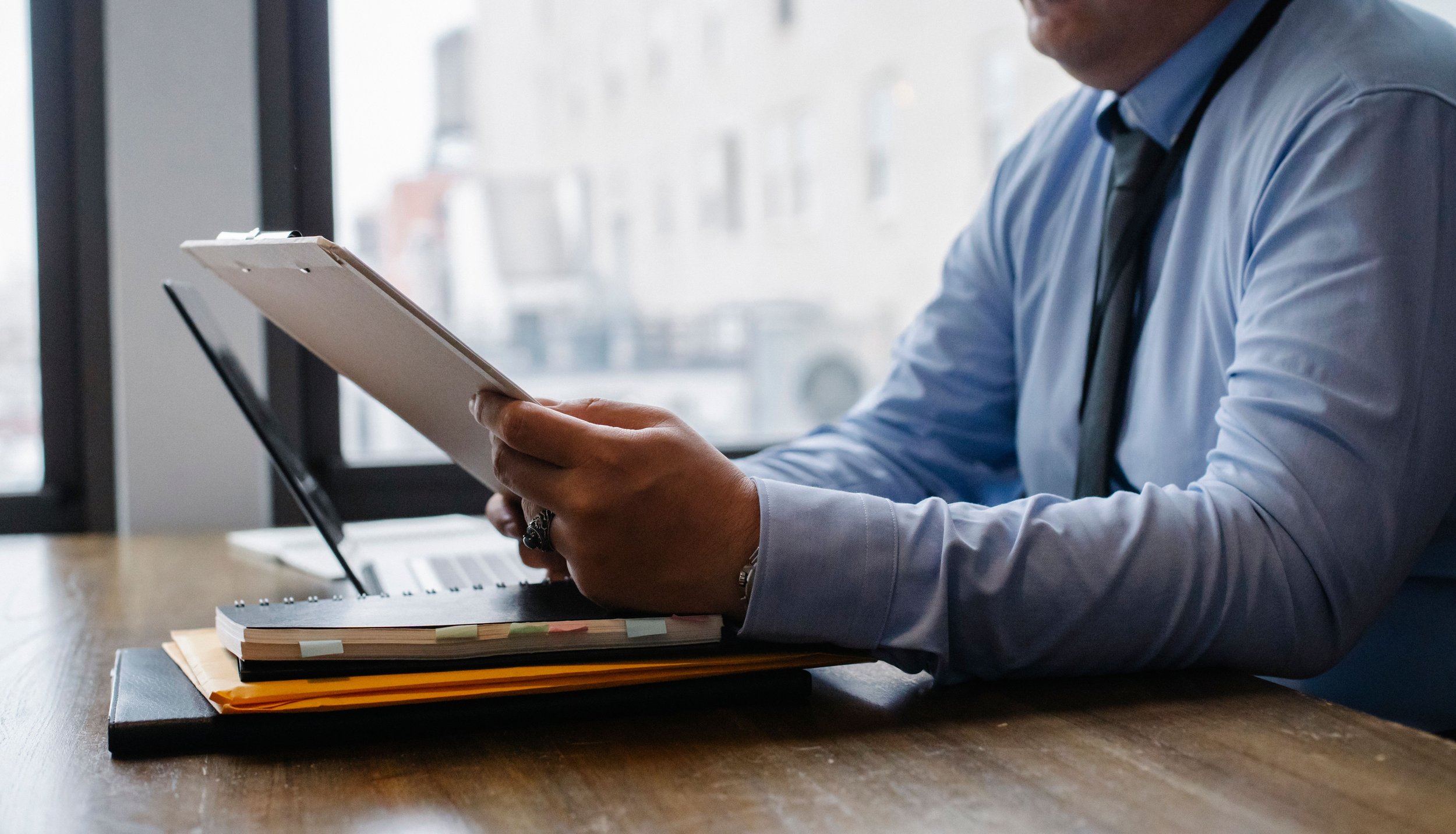 Man in a shirt and tie reviewing a document at a desk by an office window