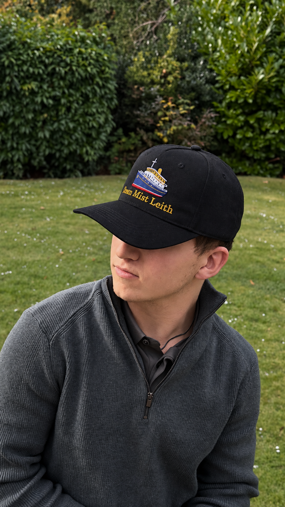 Young man wearing a black cap with embroidered boat and text, outside on green grass with bushes in background.